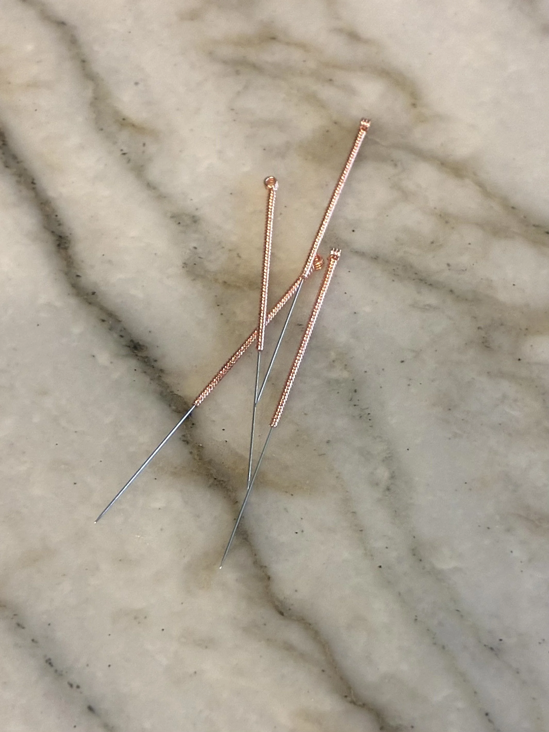 Several rose gold sewing needles with textured shafts on a marble surface.