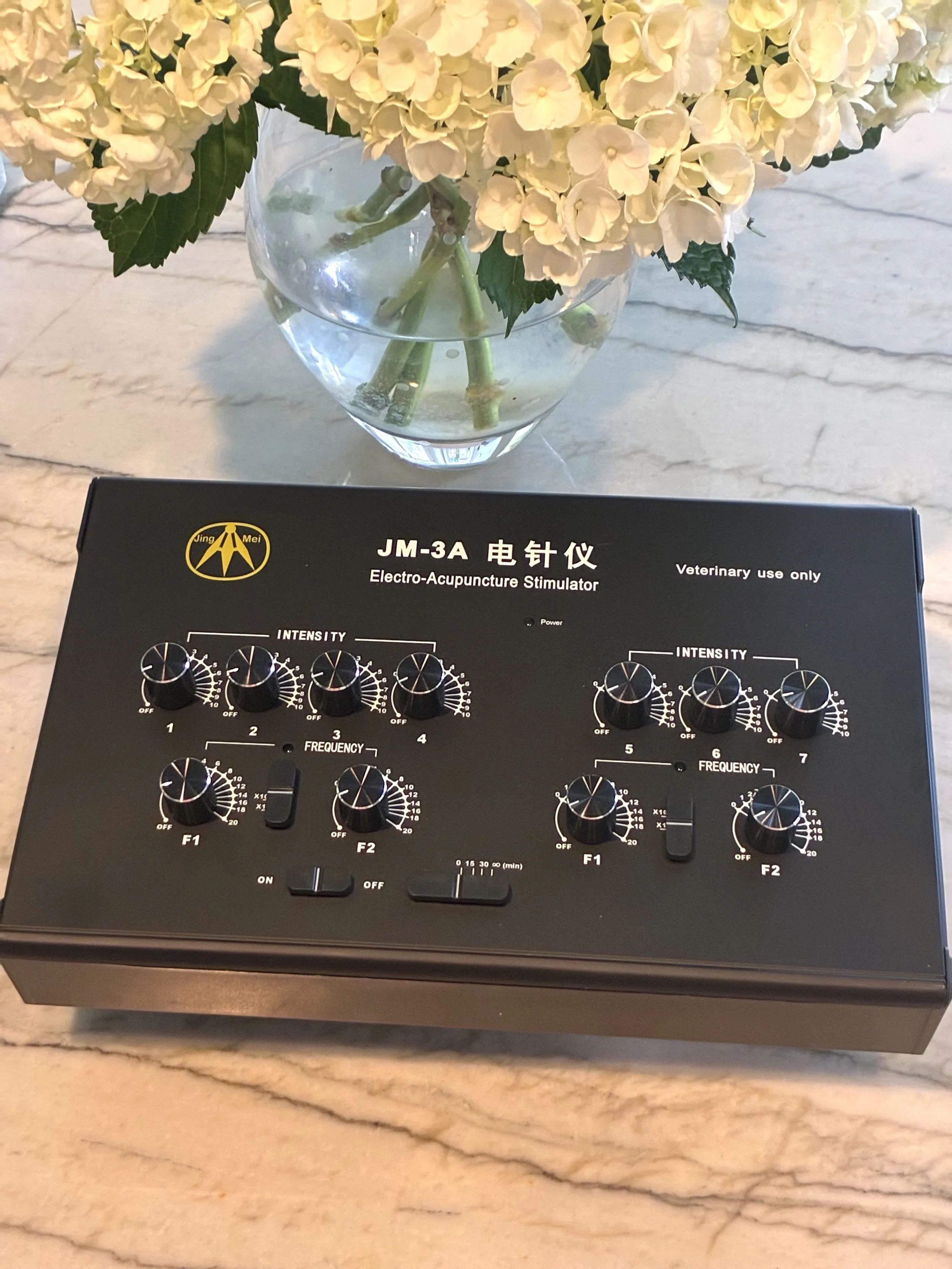 A black electro-acupuncture stimulator device labeled 'JM-3A' rests on a marble surface. It has multiple knobs for adjusting intensity and frequency, and a power switch.