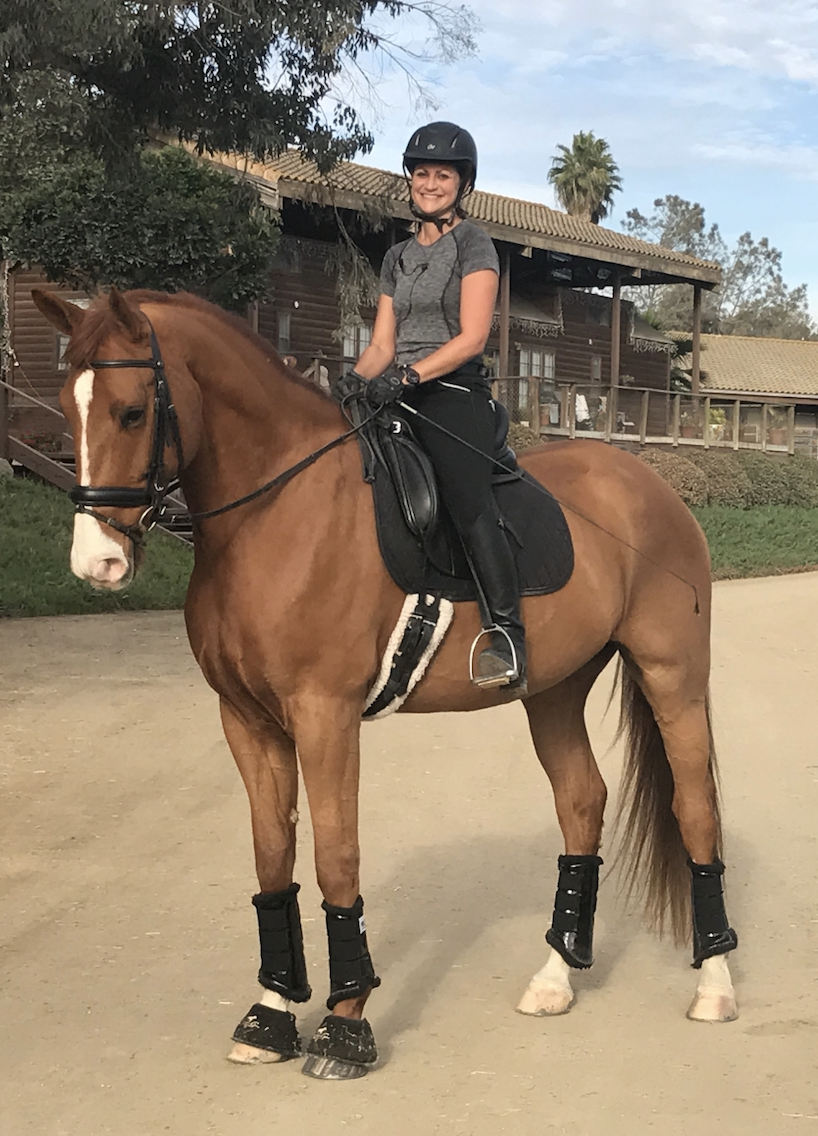 A woman smiling while riding a brown horse with white markings on its face, wearing a black helmet, gray T-shirt, black riding pants, and protective leg gear, on a dirt path in a park or farm setting with trees and buildings in the background.