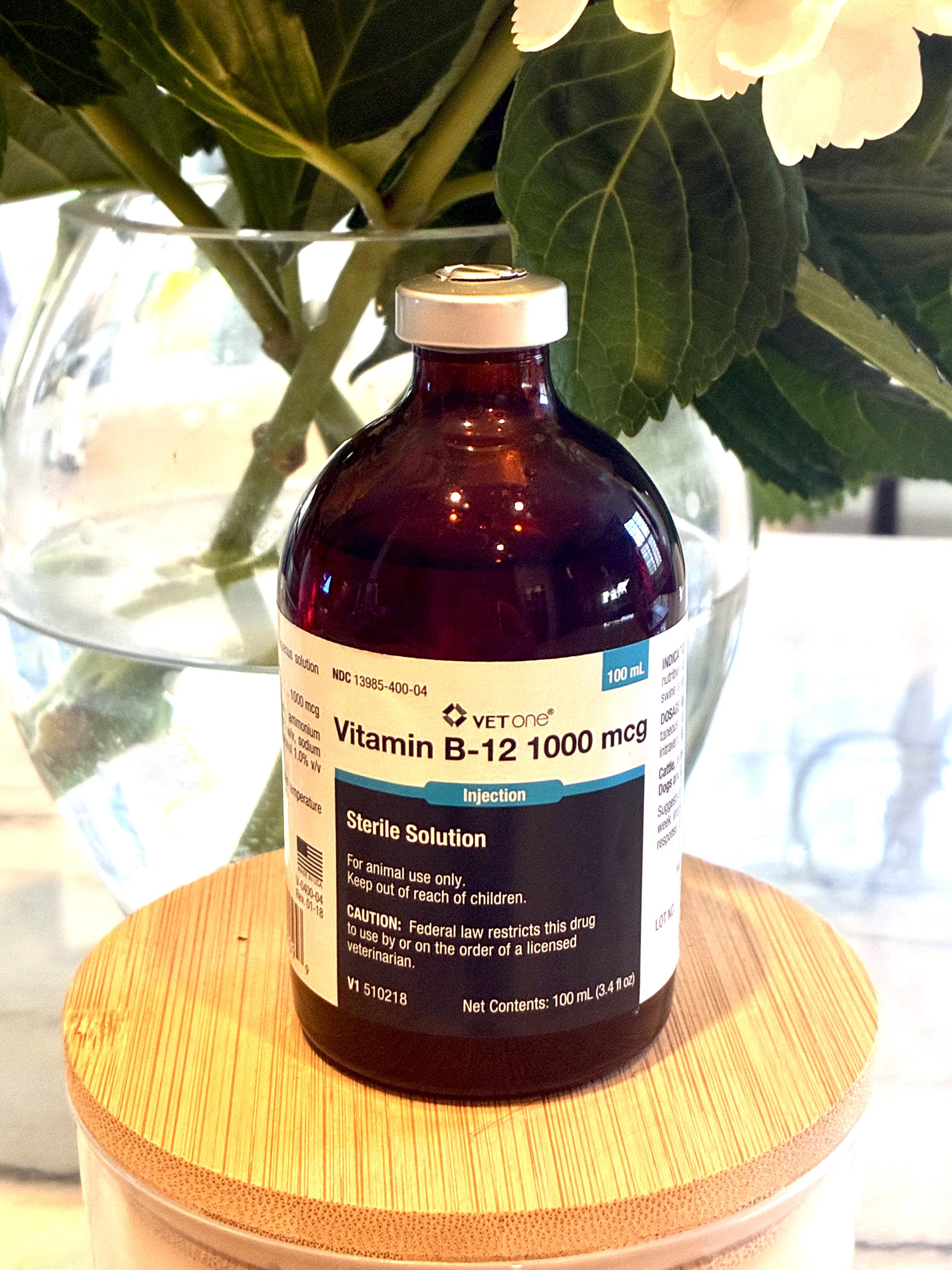 A bottle of VetOne Vitamin B-12 1000 mcg injection placed on a round, wooden table with a floral arrangement in a glass vase in the background.