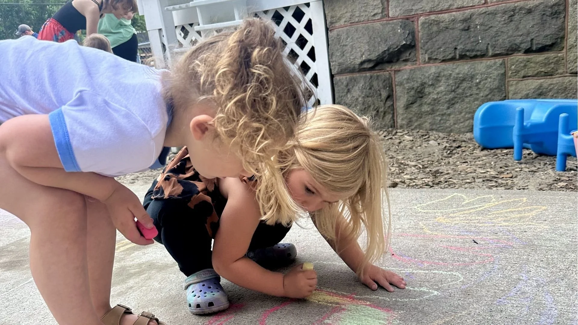 Two young children, a boy with curly blonde hair and a girl with straight blonde hair, drawing with chalk on the sidewalk at a park or playground. There are other children and adults in the background, and a small blue plastic toy lying on the ground near a brick wall.