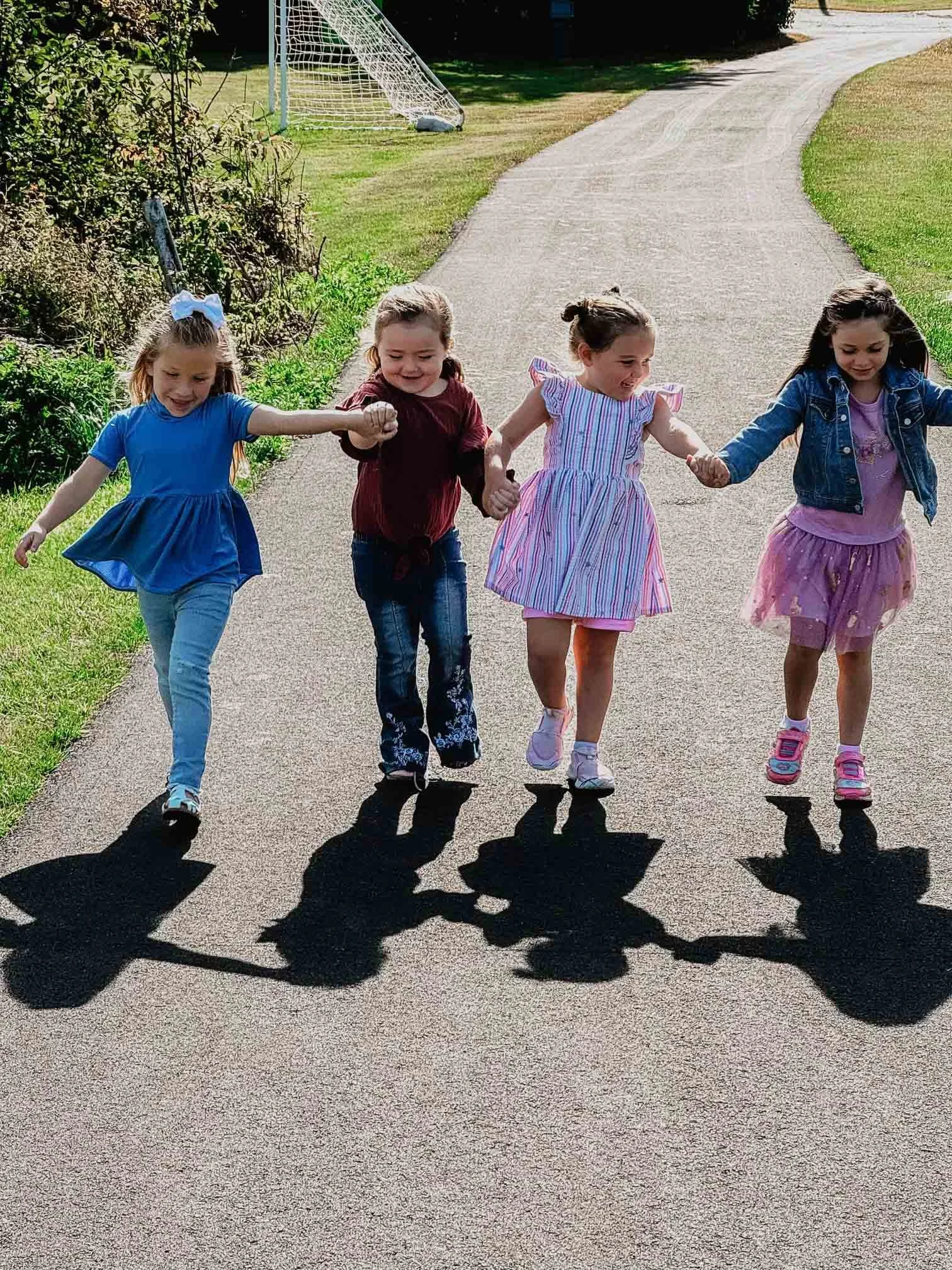 Four young girls holding hands and walking on a paved road with their shadows cast on the ground. A soccer goal and greenery are in the background.