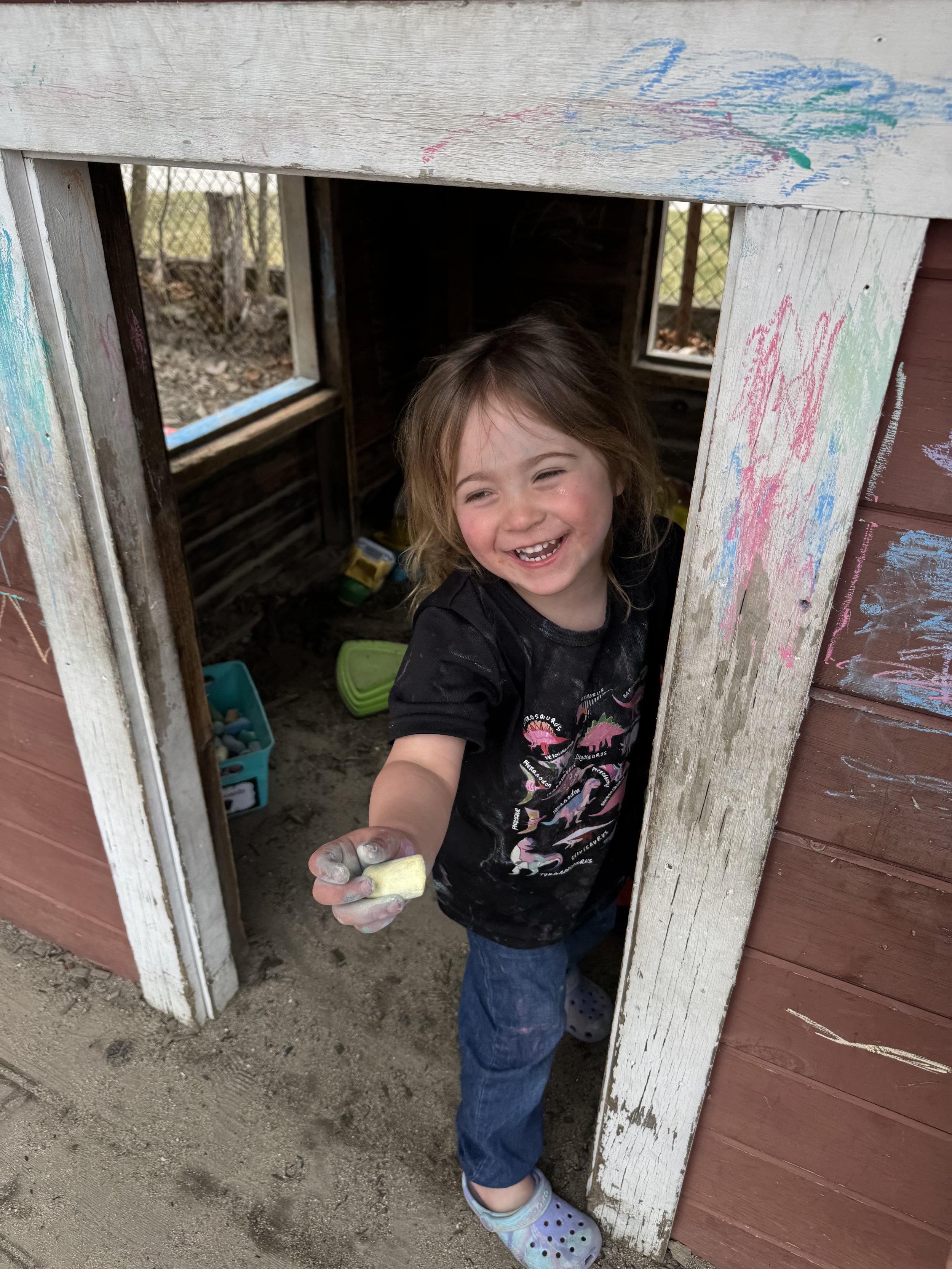 A young girl with messy hair, smiling and holding a small yellow block, standing inside a small wooden playhouse with colorful chalk drawings on the outside frame.
