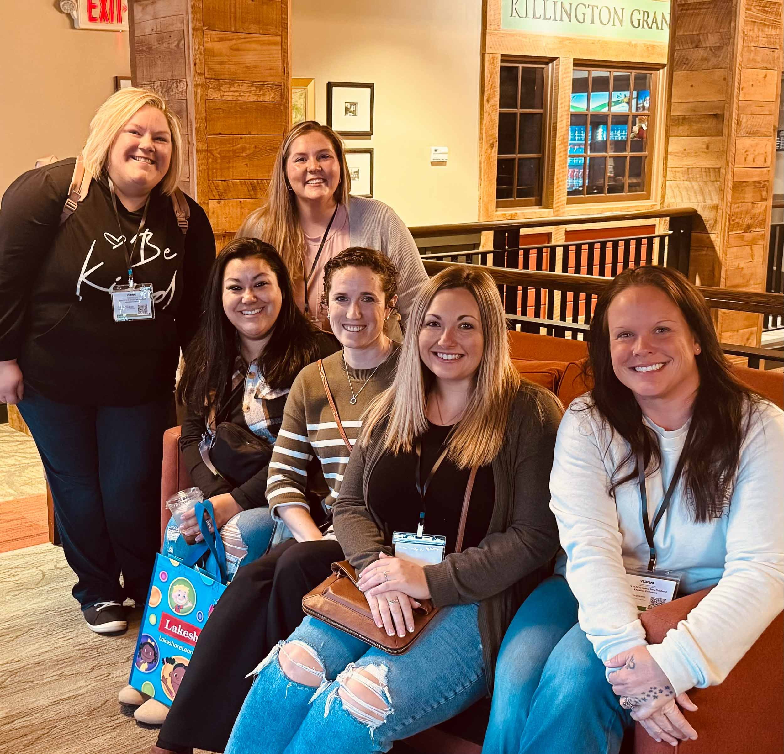 Group of women posing on a blue bench inside a wooden-paneled room, with a large window and a sign that reads 'KILLINGTON GRA'. Some women are smiling and wearing lanyards, with one holding a blue bag and another with a brown purse.