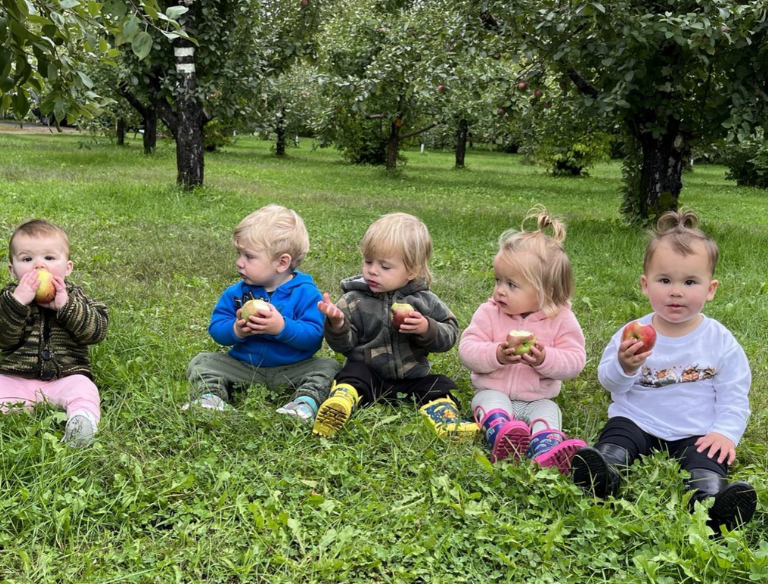 Five young children sitting on grass in an orchard, eating apples with trees and greenery in the background.