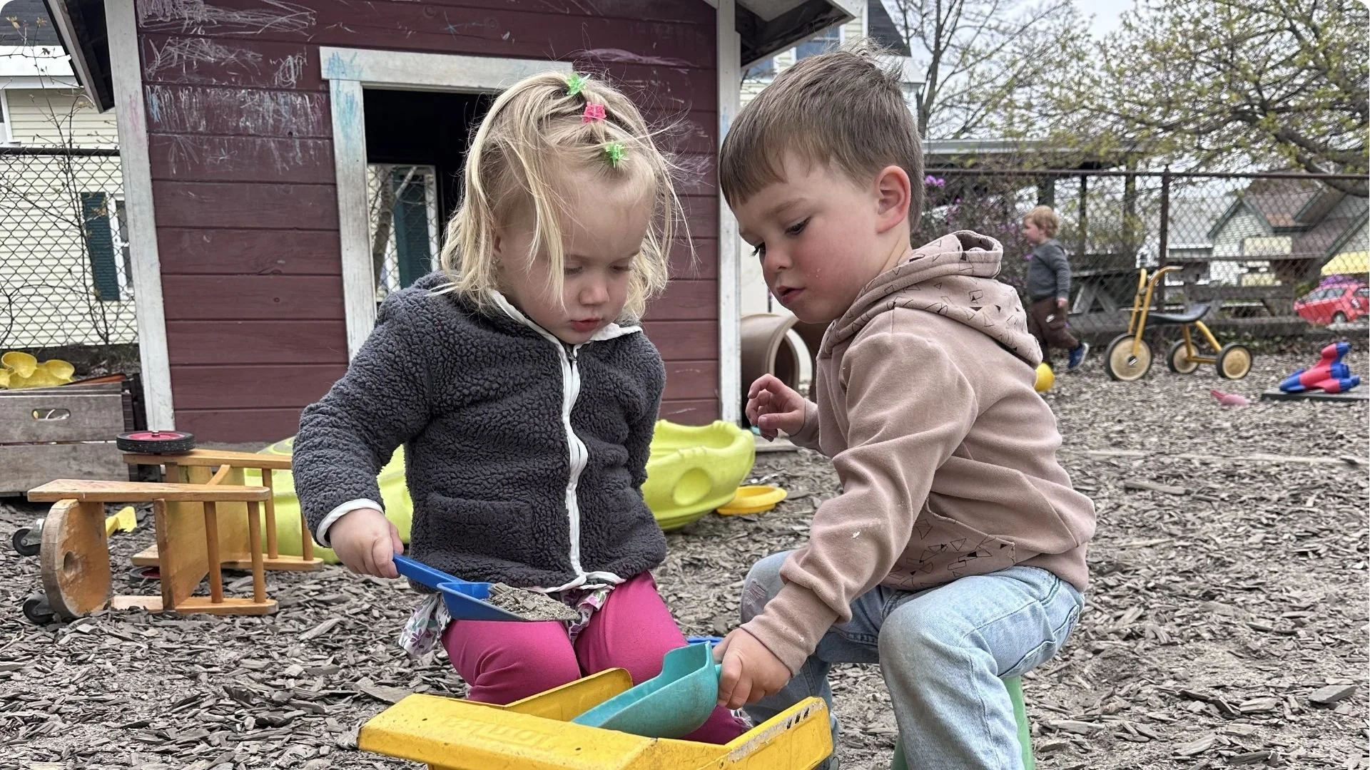 Two children are playing together on a dirt ground covered with wood chips in a backyard. They are focused on a yellow toy truck. In the background, there is a red wooden playhouse, a fence, a tricycle, and other toys.