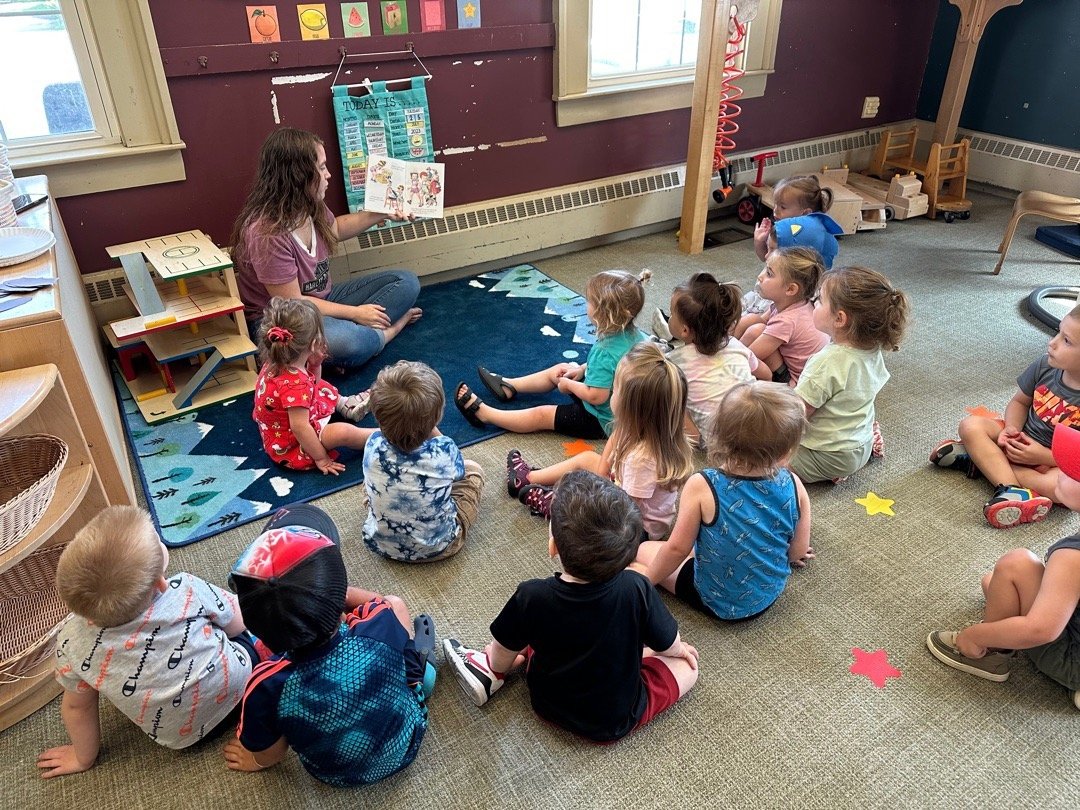 A young woman is sitting cross-legged on a small rug, reading a picture book to a group of children in a classroom. The children are sitting on the floor, attentively listening.