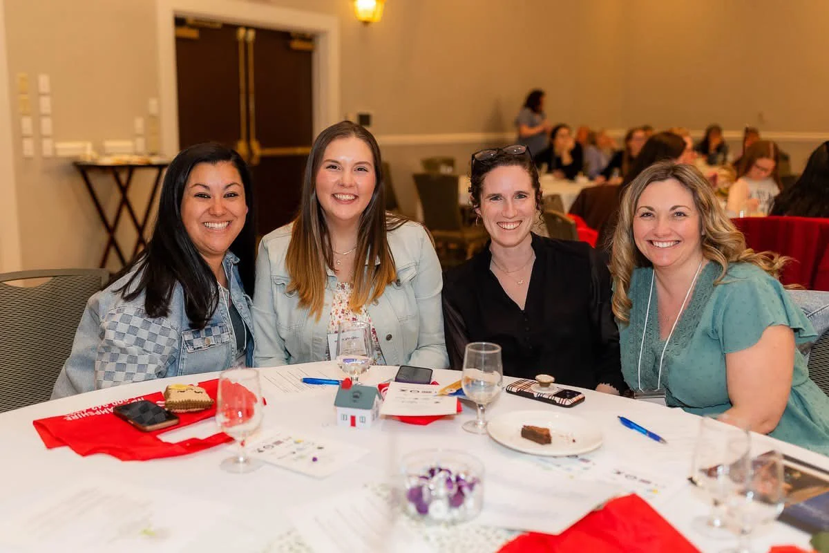 Four women sitting at a round table smiling at the camera during a social event or gathering.