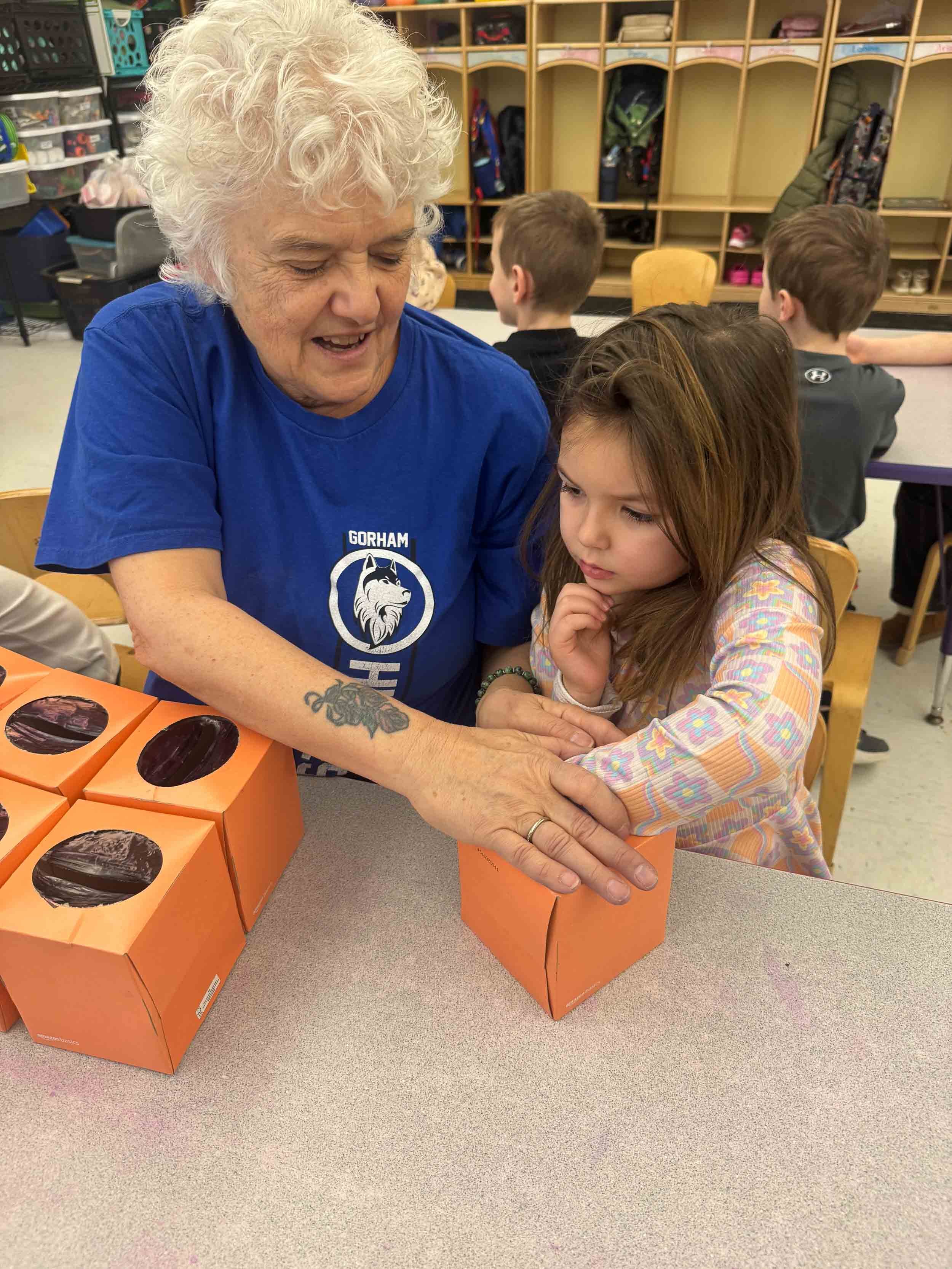 An elderly woman and a young girl are working together at a table in a classroom, placing their hands on orange boxes with black circular designs. There are other children sitting at the table in the background, with classroom cubbies and shelves visible.