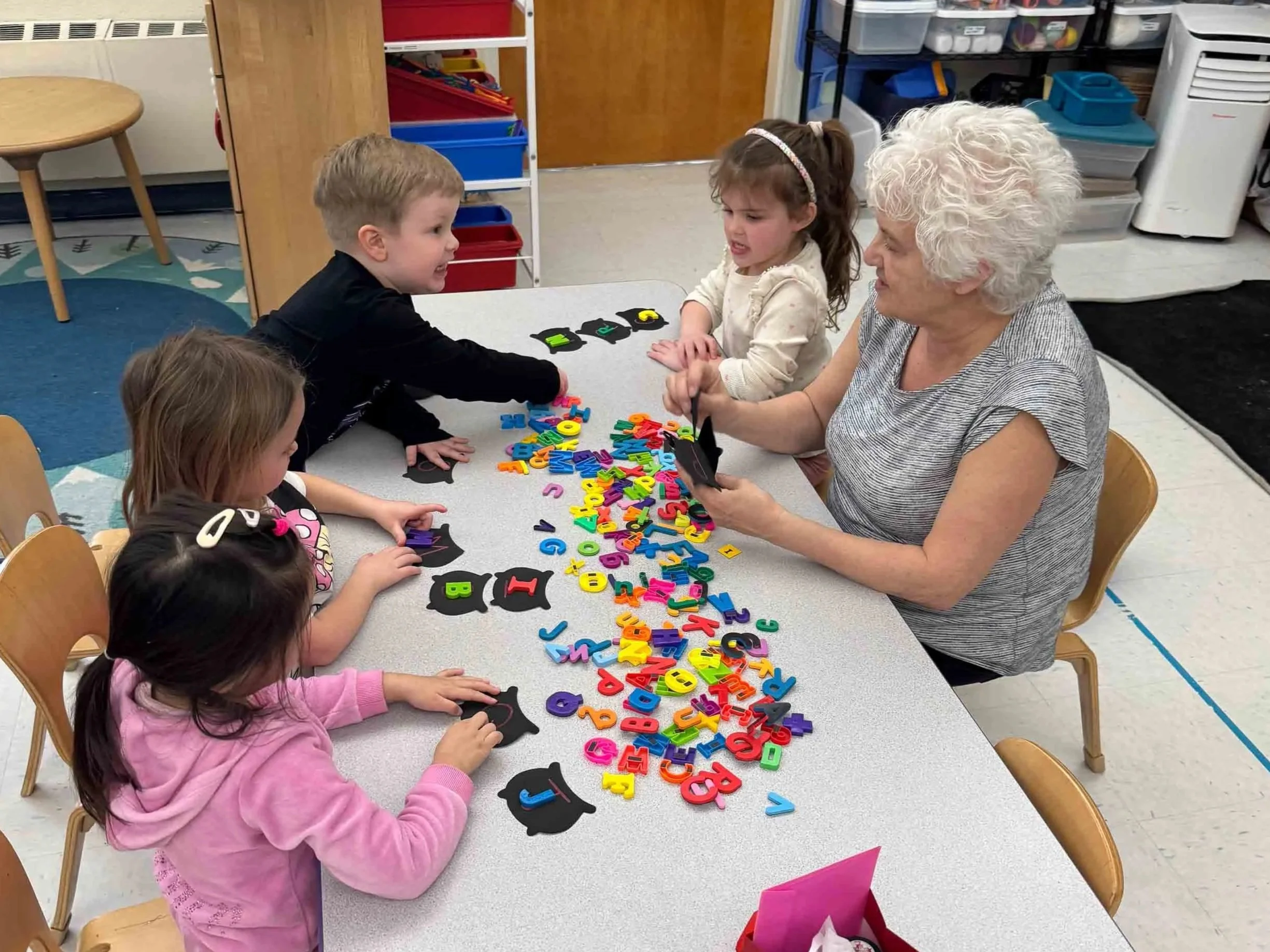 A group of young children sitting around tables with colorful alphabet letters and black chalkboard cutouts, engaging in an educational activity with an older woman.