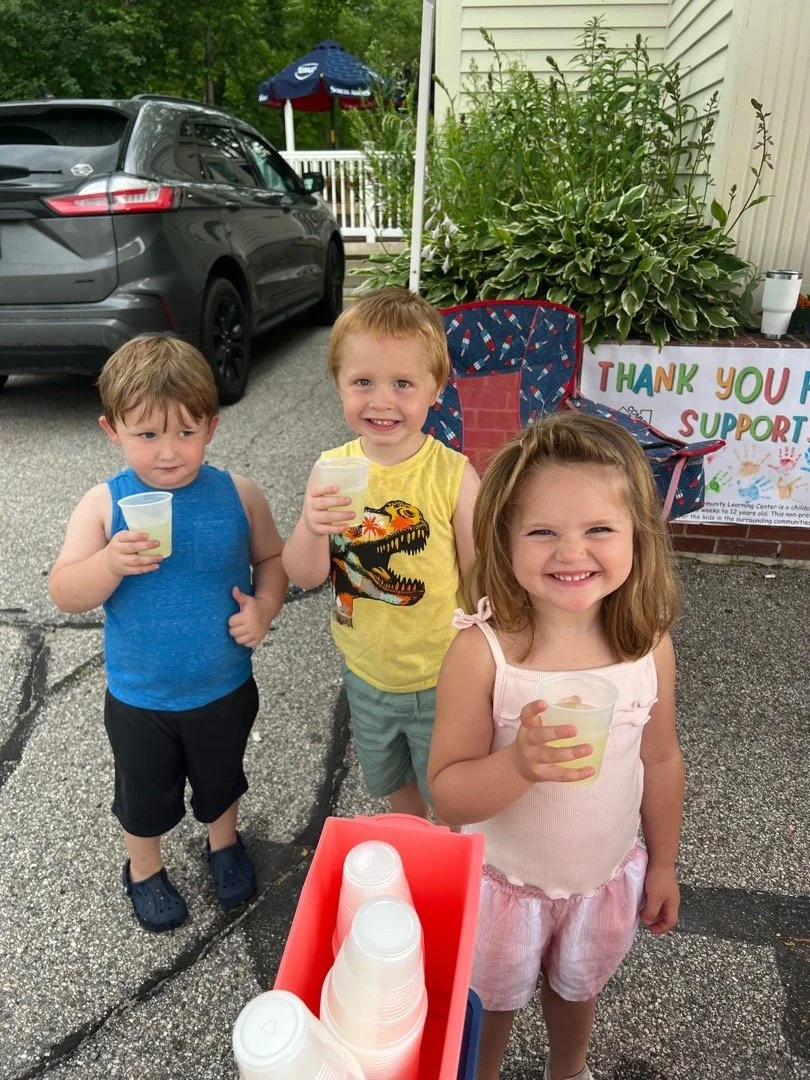 Three young children standing outdoors with cups of lemonade, smiling and enjoying a summer day.