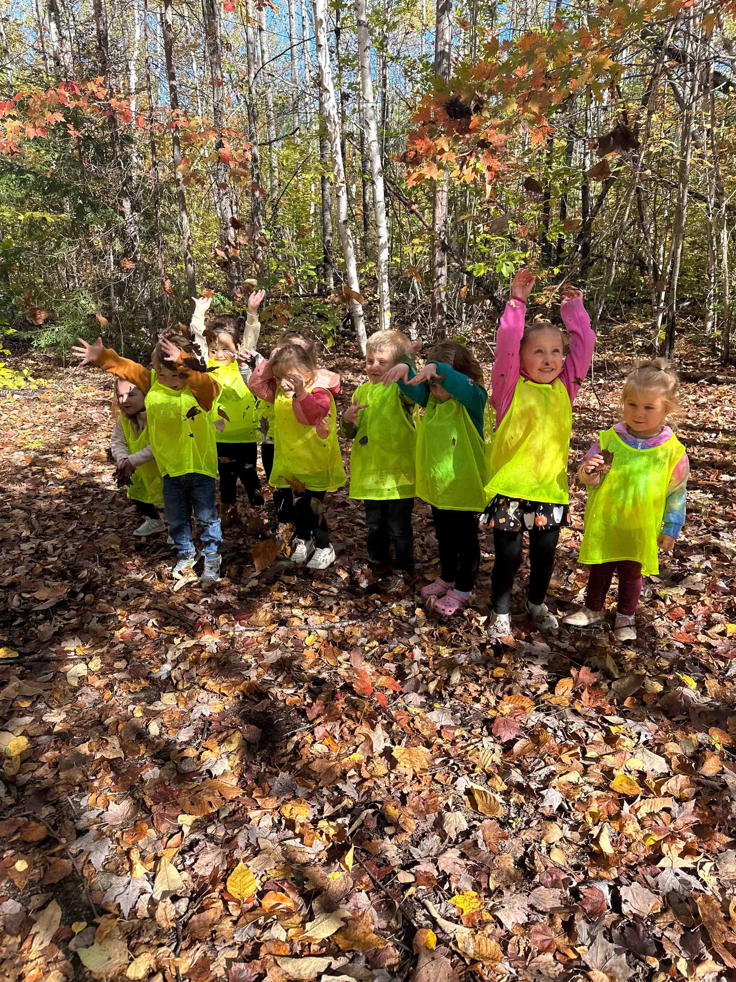 A group of children wearing bright yellow vests playing in an autumn forest, surrounded by fallen leaves and trees with colorful foliage.
