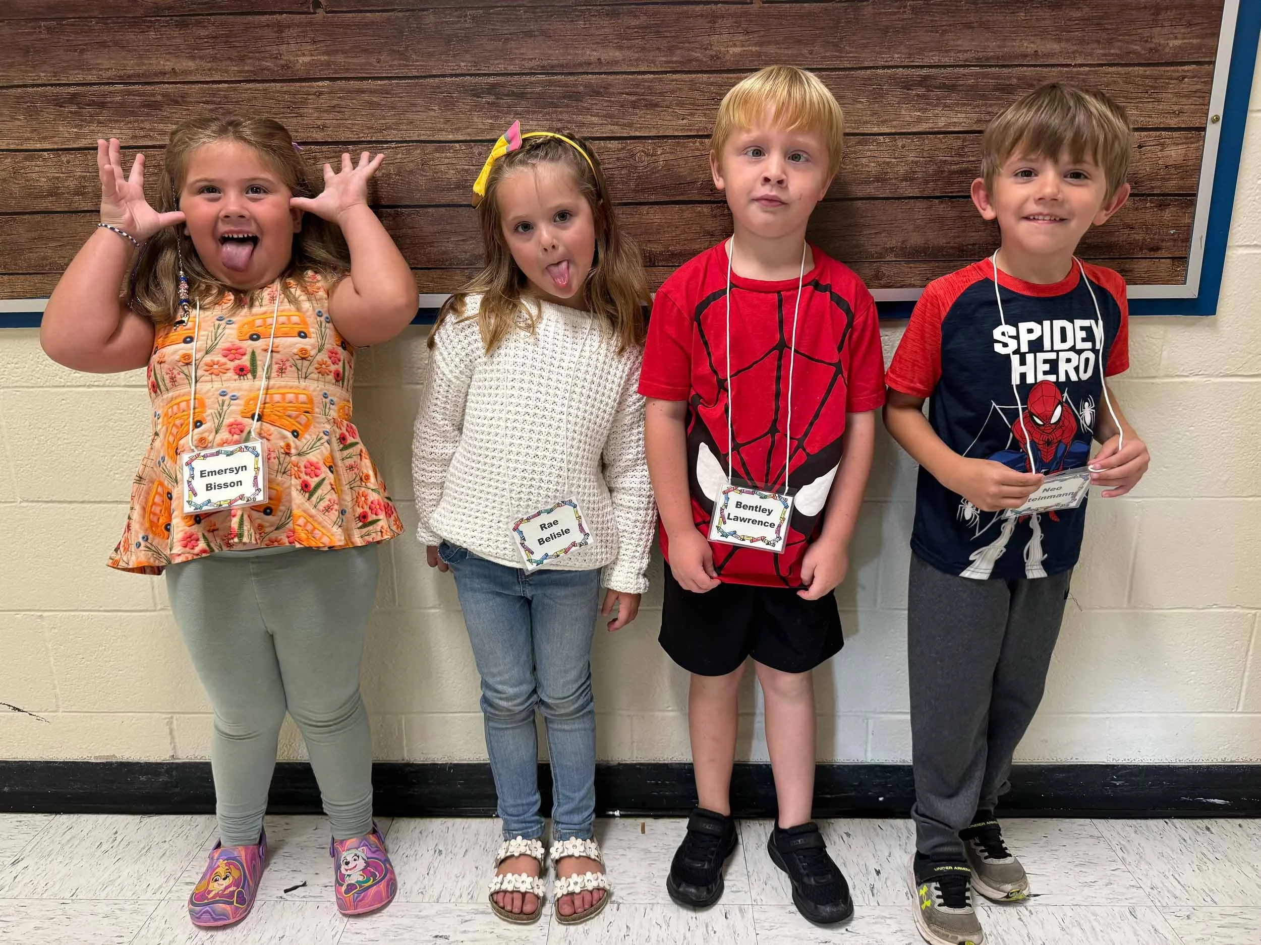 Four children standing in front of a wooden wall, making playful faces. The first girl on the left is wearing a floral dress and colorful shoes, sticking out her tongue and holding her hands near her face. The second girl is wearing a white sweater and jeans, also sticking out her tongue. The third child, a boy, is wearing a red superhero T-shirt with a spider web design and black shorts, looking serious. The fourth boy is wearing a navy and red shirt with a Spider-Man logo and gray pants, smiling.