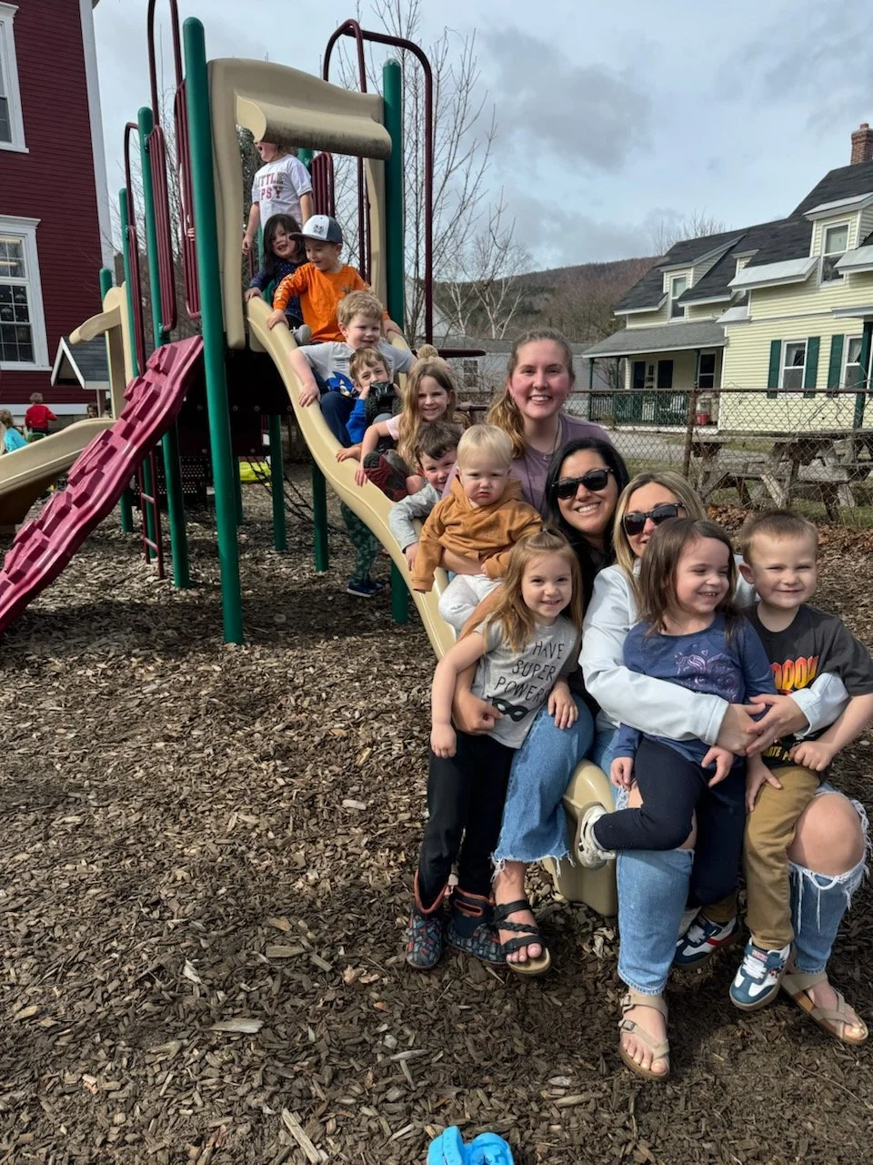 Group of children and adults sitting on a slide at a playground, smiling and enjoying outdoor play.