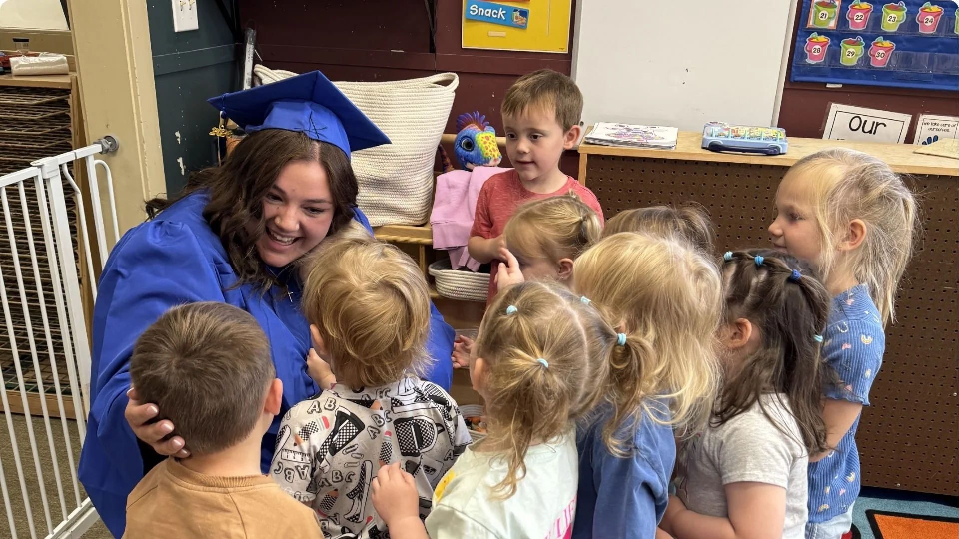 A group of young children gathered around a woman in a blue cap and gown. The children are listening and engaging with her, with some looking up at her and others interacting with each other.
