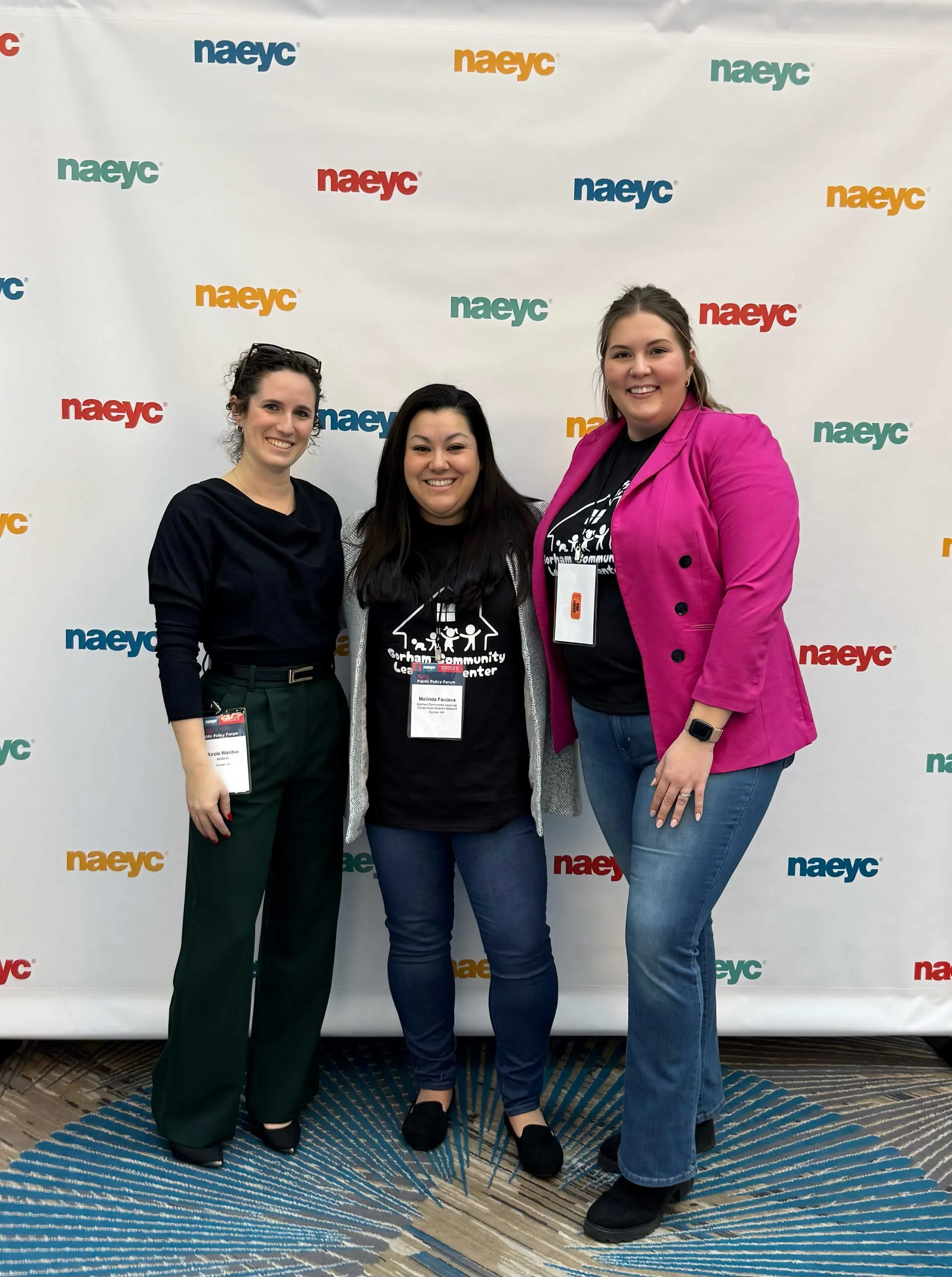Three women standing in front of a NAEYC branded backdrop at an event, smiling at the camera. The woman on the left has curly hair and is wearing a black top with green pants. The woman in the middle has long dark hair and is wearing a black event badge. The woman on the right is taller, has light brown hair, and is wearing a bright pink blazer and jeans.