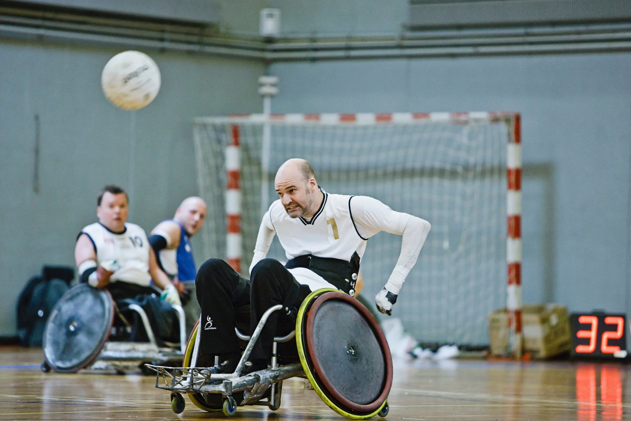 A guy in a wheelchair playing wheelchair rugby in an indoor gymnasium, with a goalpost and a digital score display showing 32 points, and two other players in wheelchairs in the background.