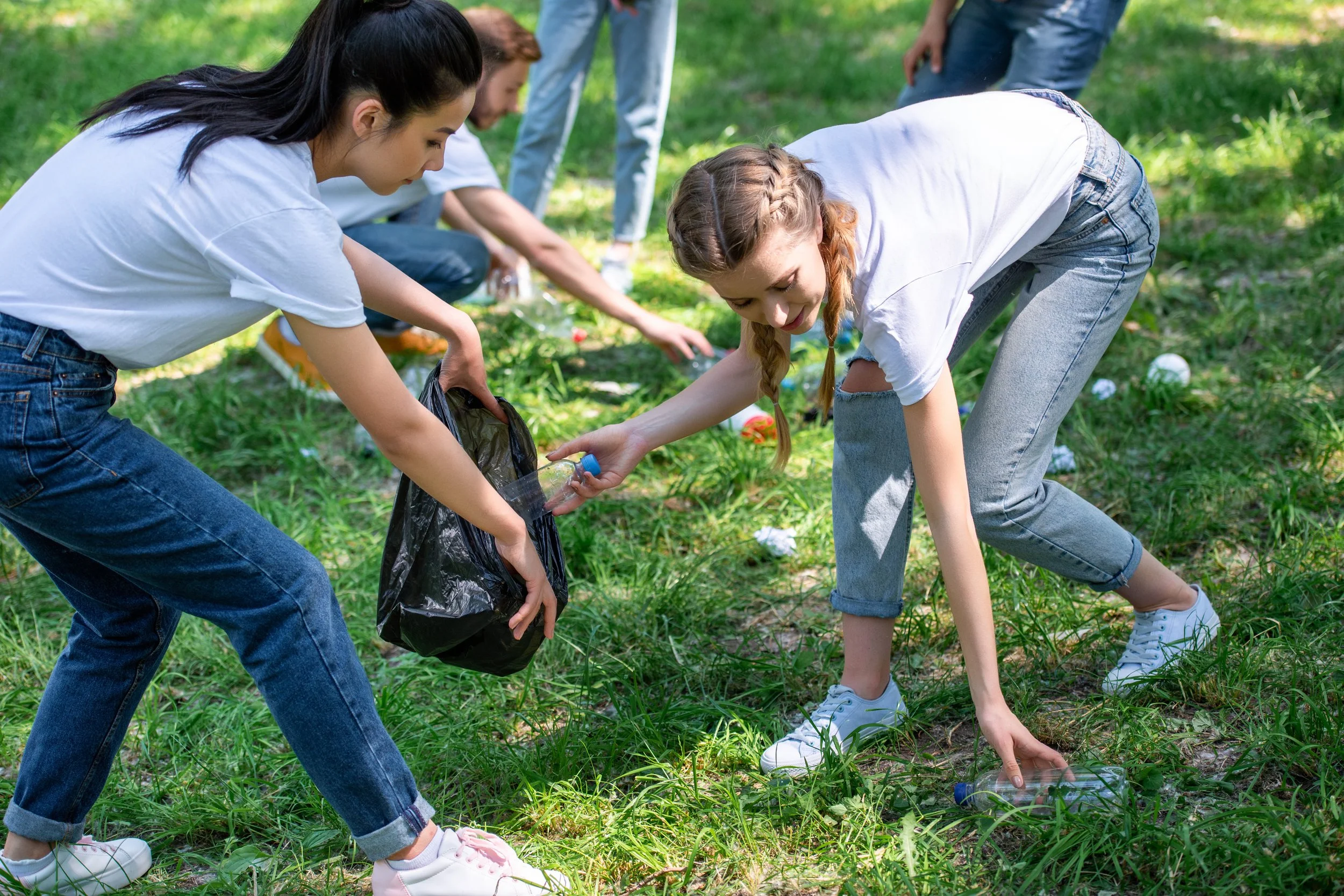 Young girls participating in a cleanup activity in a grassy outdoor area, collecting litter and placing it into trash bags.
