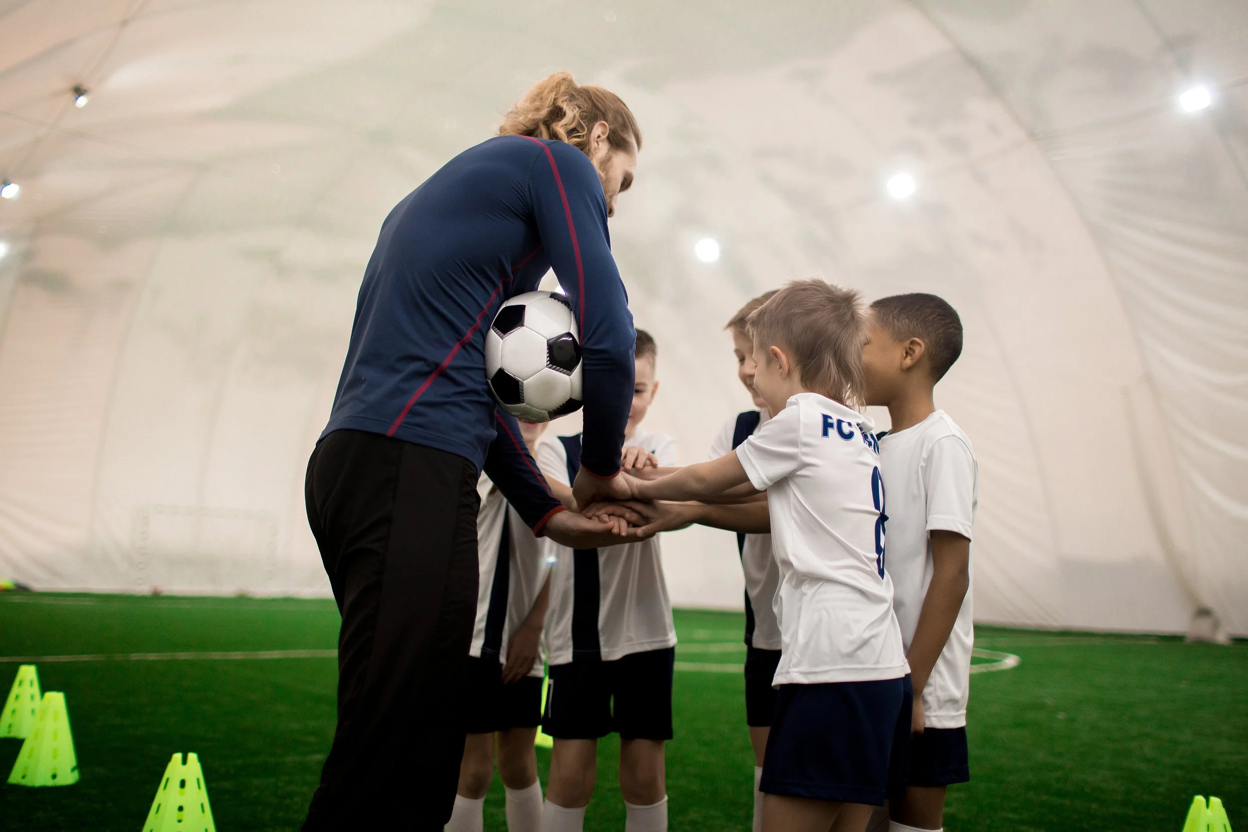 A coach with long hair, wearing a navy jacket, holding a soccer ball, is sharing a team huddle with young girls in white soccer jerseys inside a large indoor soccer facility.