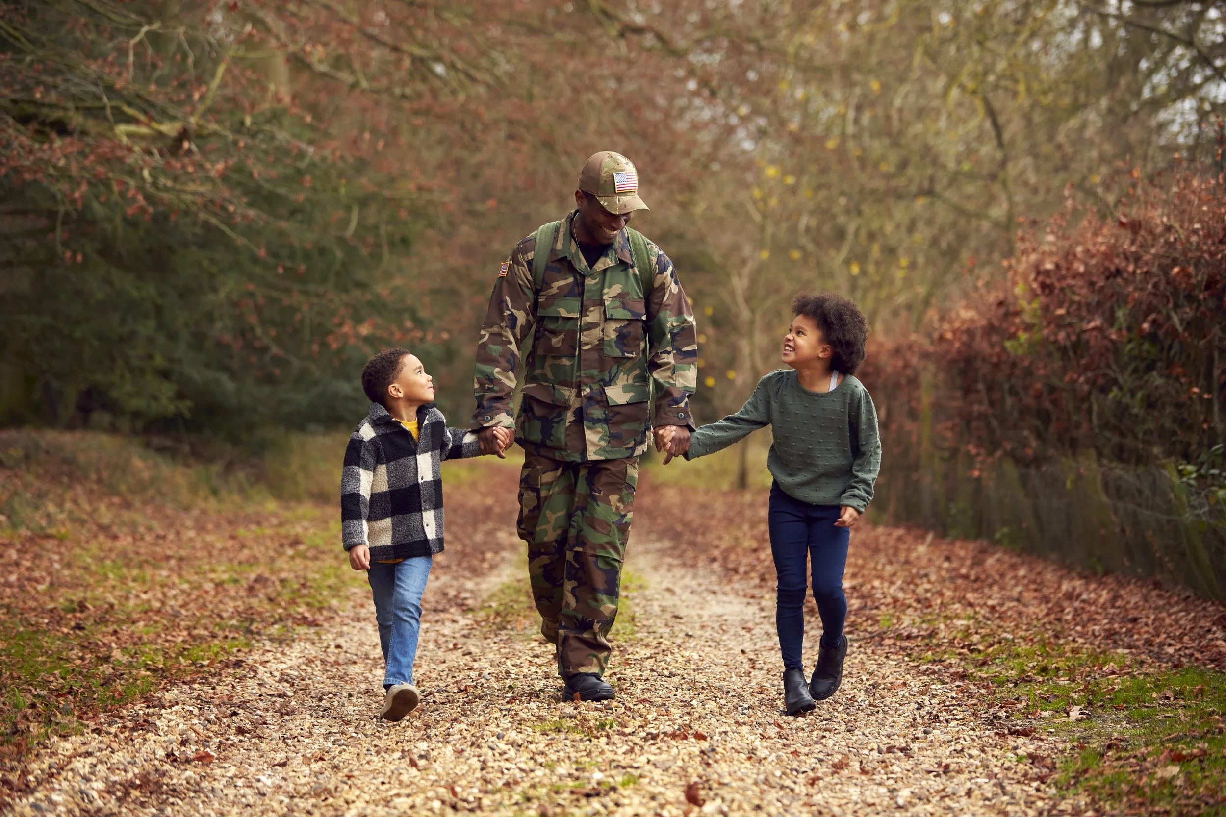 A soldier walking outdoors on a pathway with two children, one boy and one girl, holding hands and smiling in a park-like setting with fall foliage.