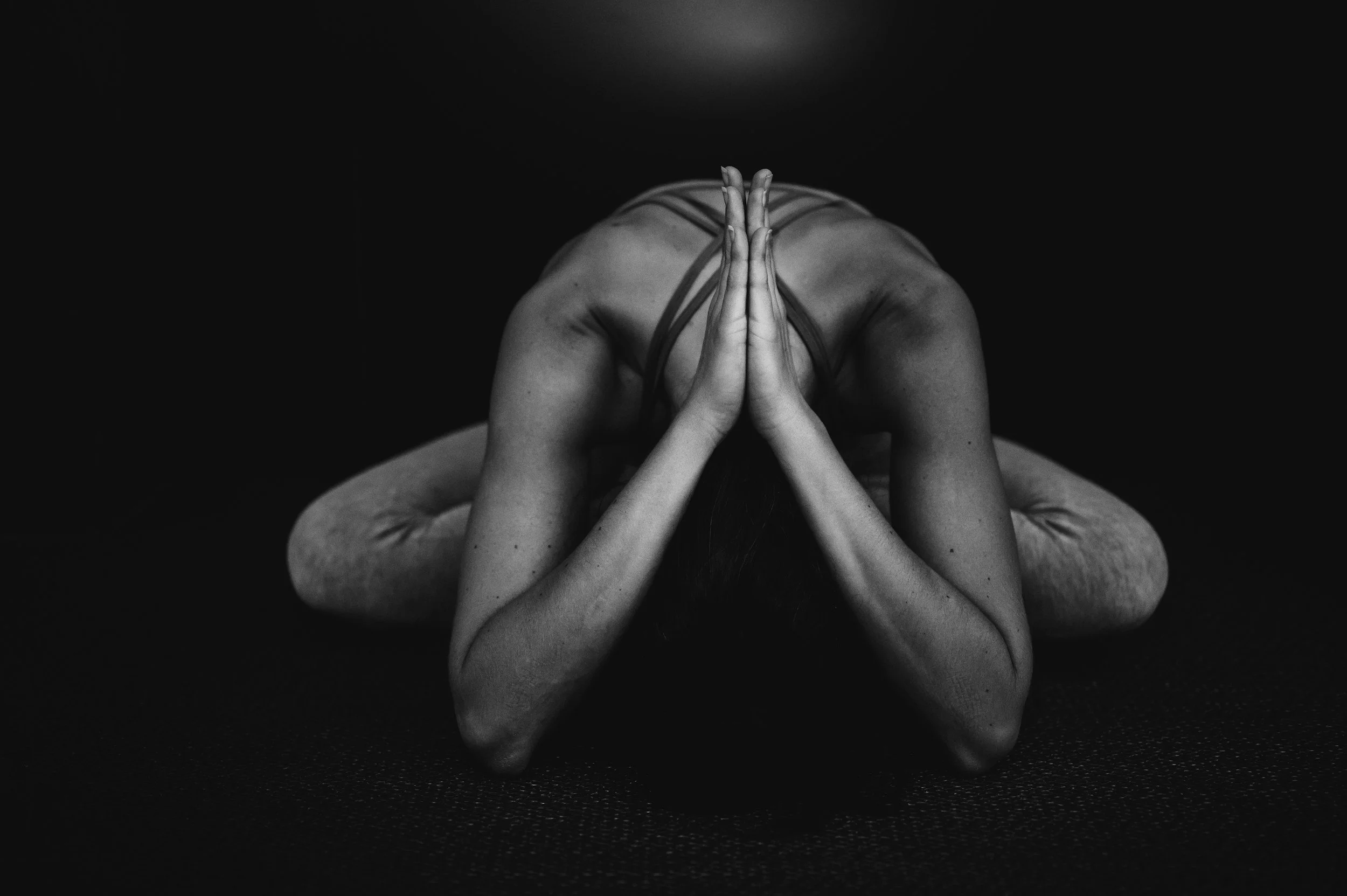 A person practicing yoga lying down with hands pressed together in a prayer position, head bowed, captured in black and white against a dark background.