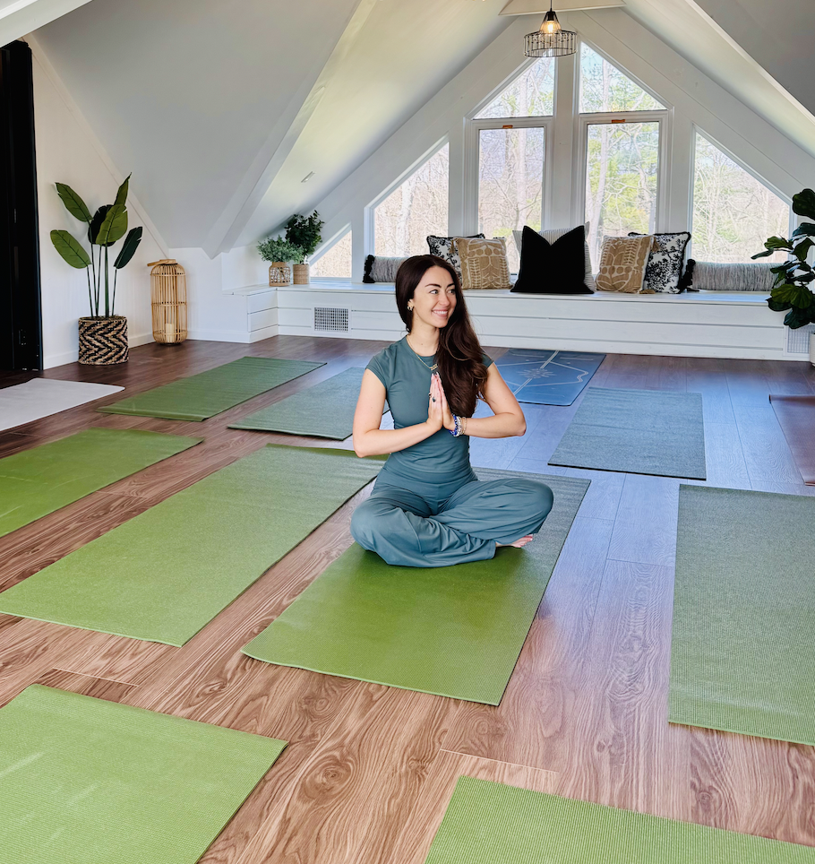 A woman practicing yoga in a bright attic room with a large window seat, green yoga mats on wooden floor, and plants, with trees visible outside.