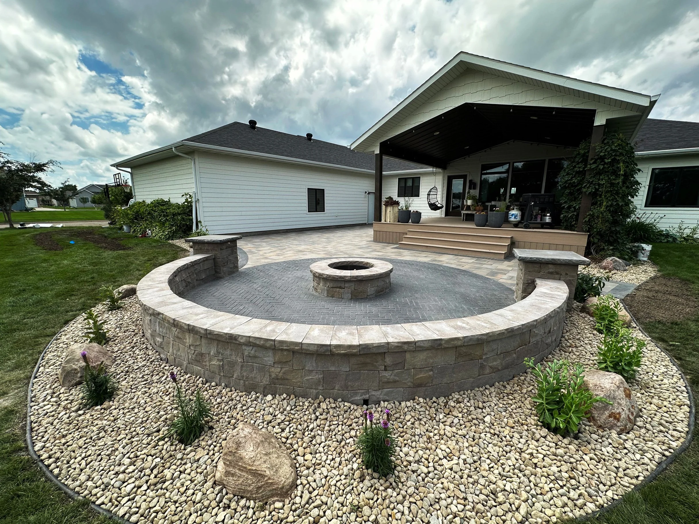 Backyard patio with circular brick fire pit, stone walls, plant bed with large rocks and flowering plants, surrounded by a grassy lawn and a house with a covered porch.