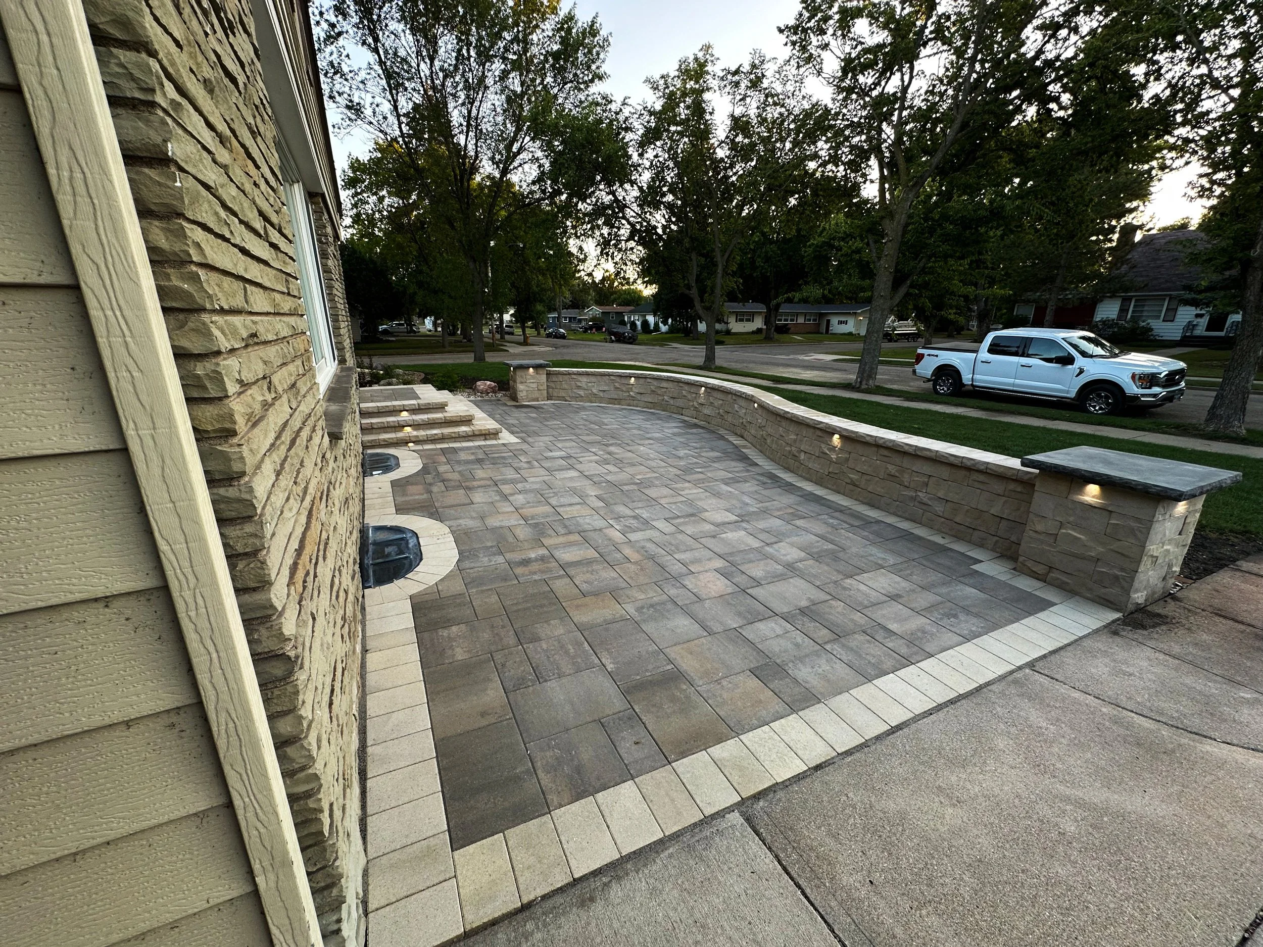 A newly paved outdoor patio area with stone tiles, a low stone wall, and built-in lighting, adjacent to a house with beige siding and stone accents, in a residential neighborhood with trees and a white pickup truck parked on the street.