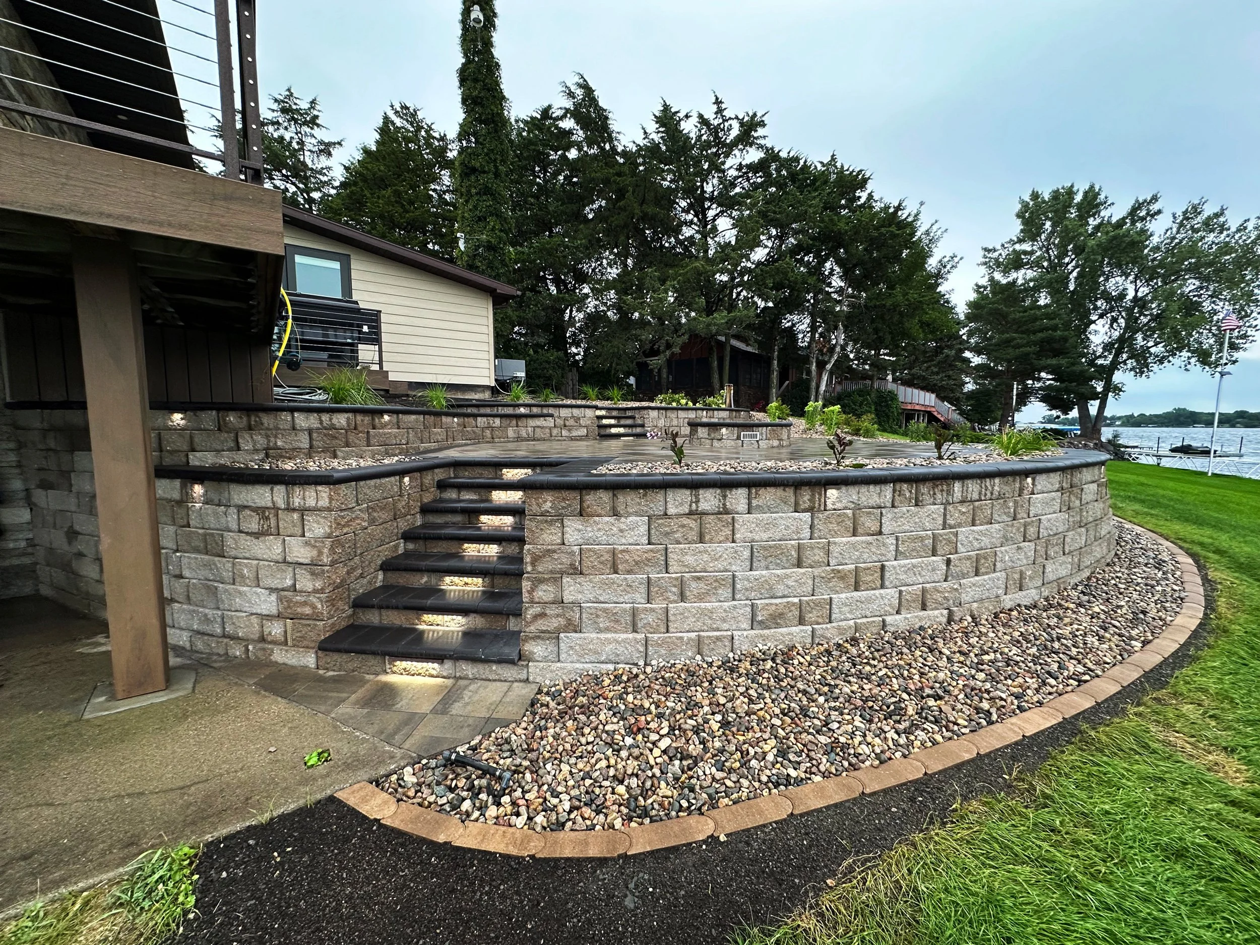 A multi-level stone patio with stairs, located next to a house with beige siding, overlooking a waterfront area with a dock and trees in the background.