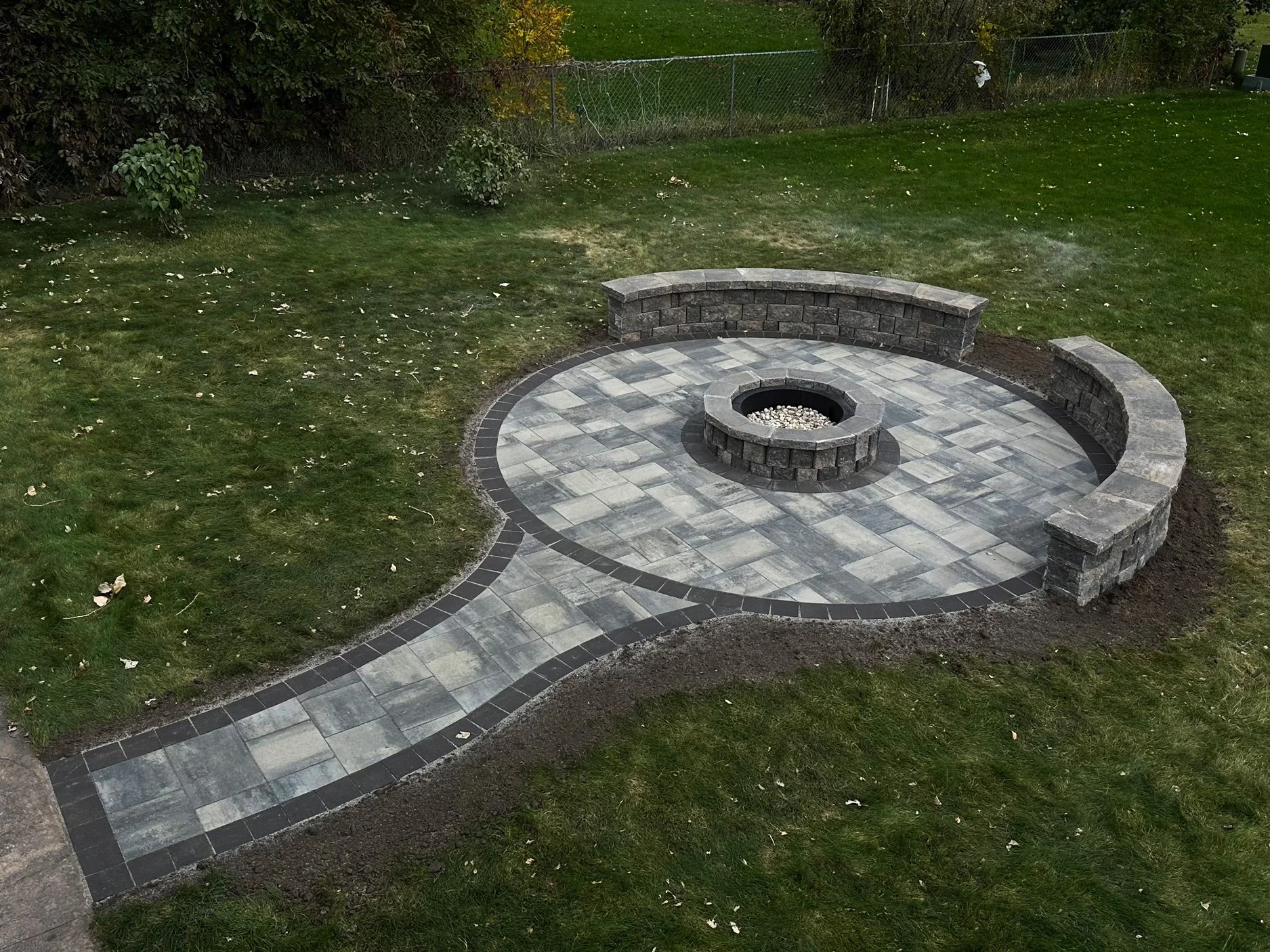 A landscaped backyard with a circular stone patio, a fire pit in the center, and built-in stone benches around the fire pit. The patio extends with a curved walkway into a grassy area with trees and a chain-link fence in the background.