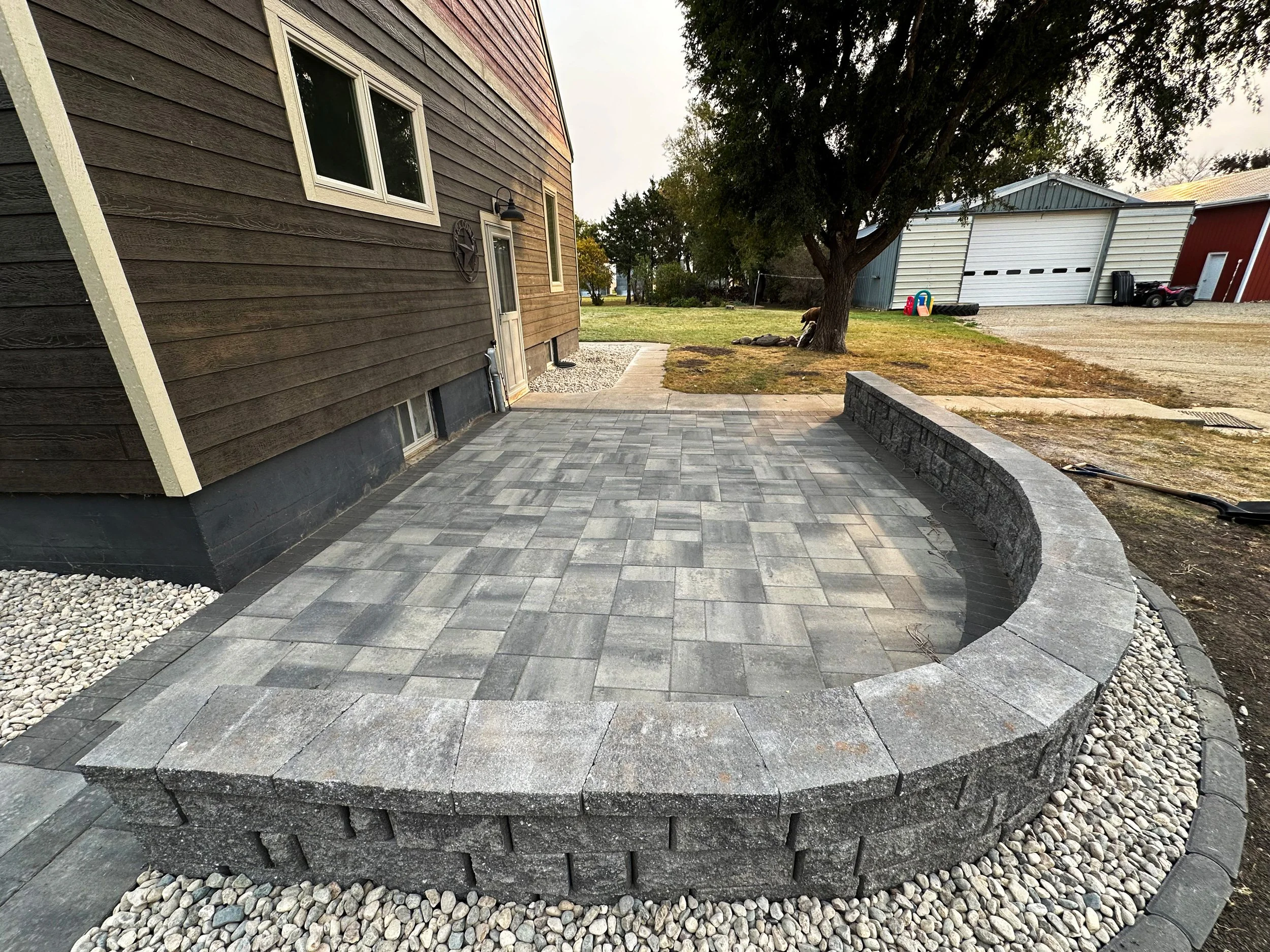A newly constructed brick patio with a curved wall in back, located next to a house with brown wooden siding and white window frames, with a tree and storage buildings in the background.