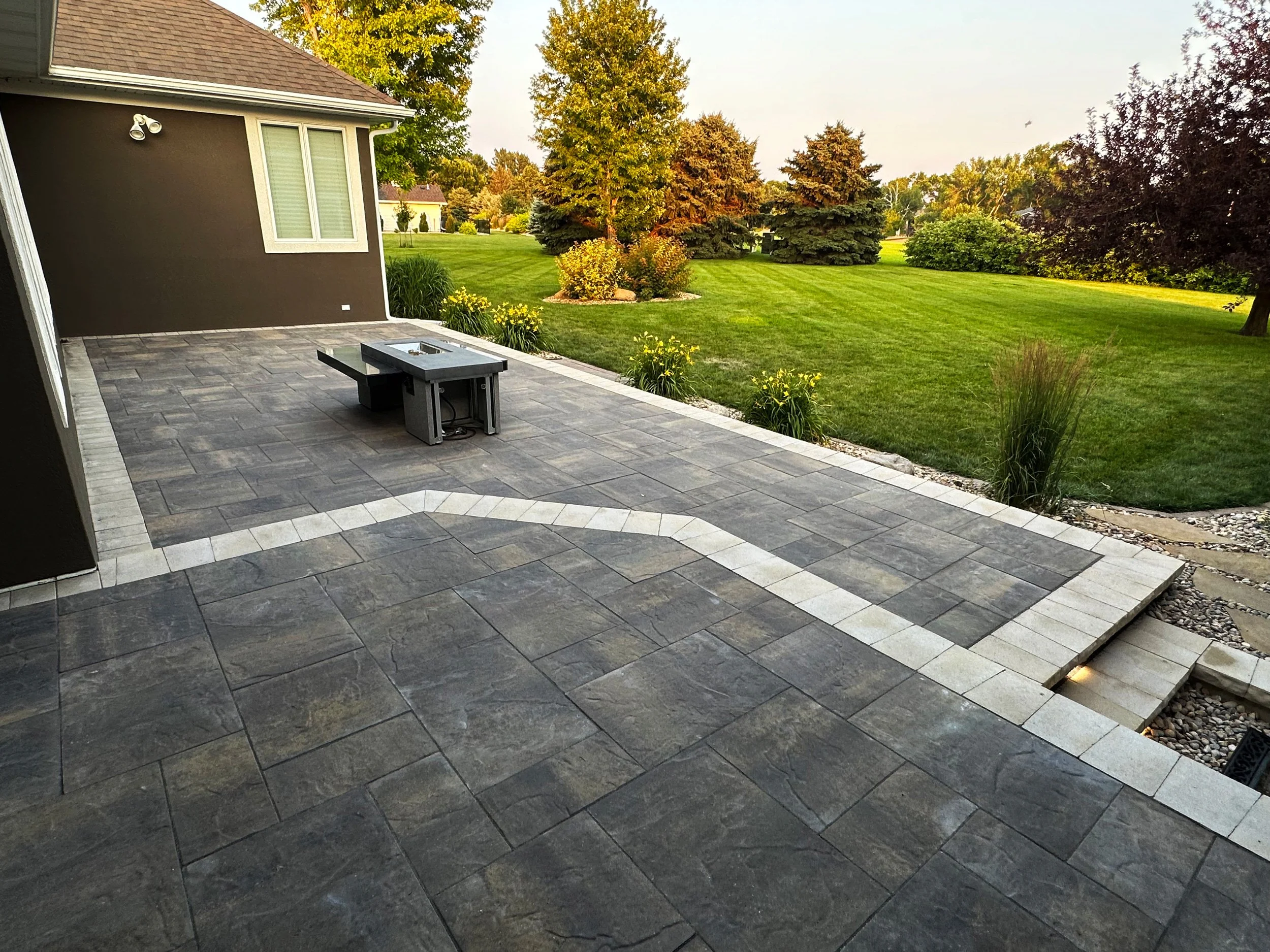 A backyard patio paved with gray and white stones, featuring a built-in fire pit and surrounded by a well-maintained green lawn with trees and shrubs.