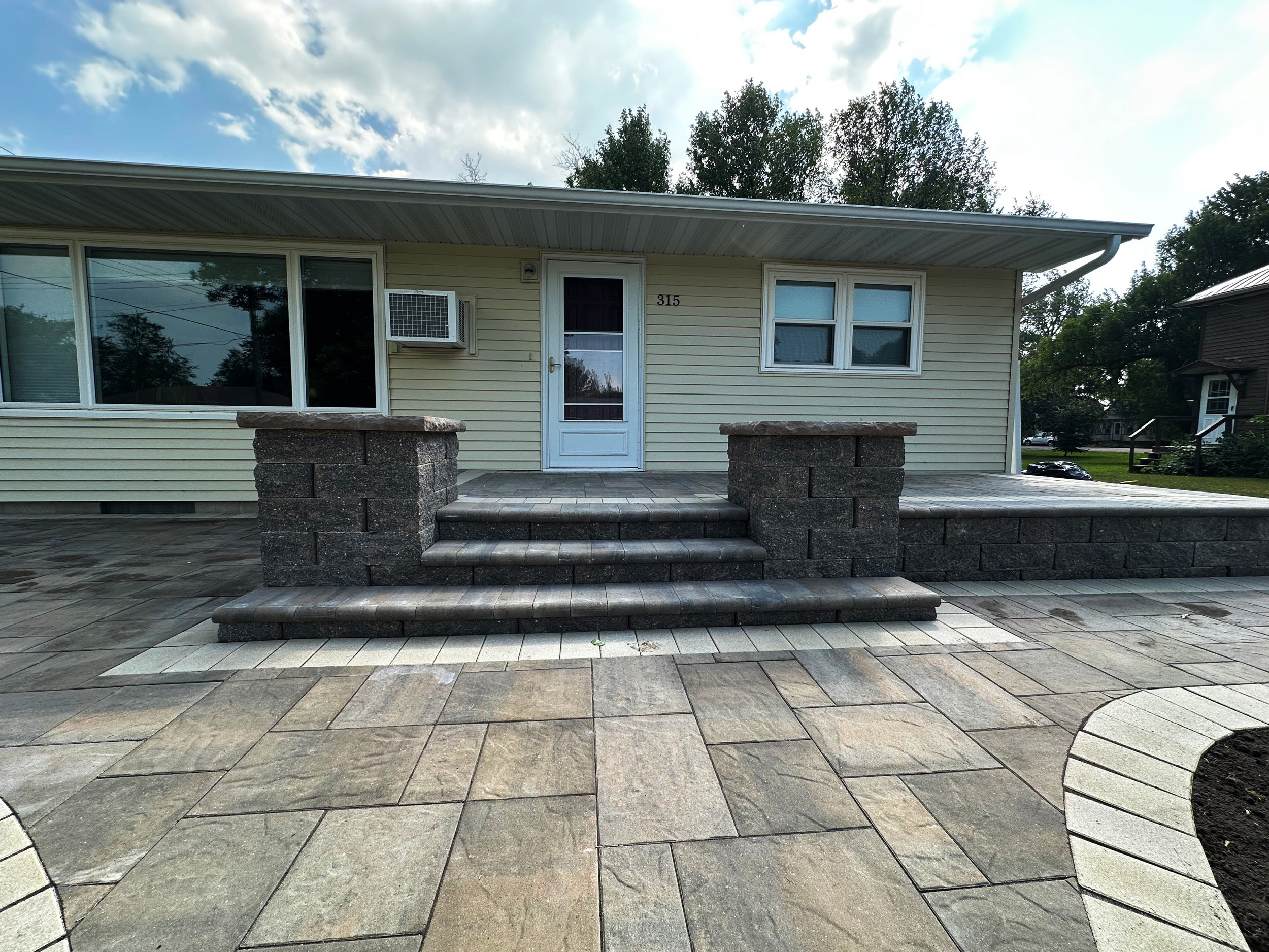 Front view of a house with beige siding, white trim, and a small front porch with stone stairs and a stone wall. The house has a window air conditioning unit, two windows, and a white door with the number 315. The yard features a large stone patio wi