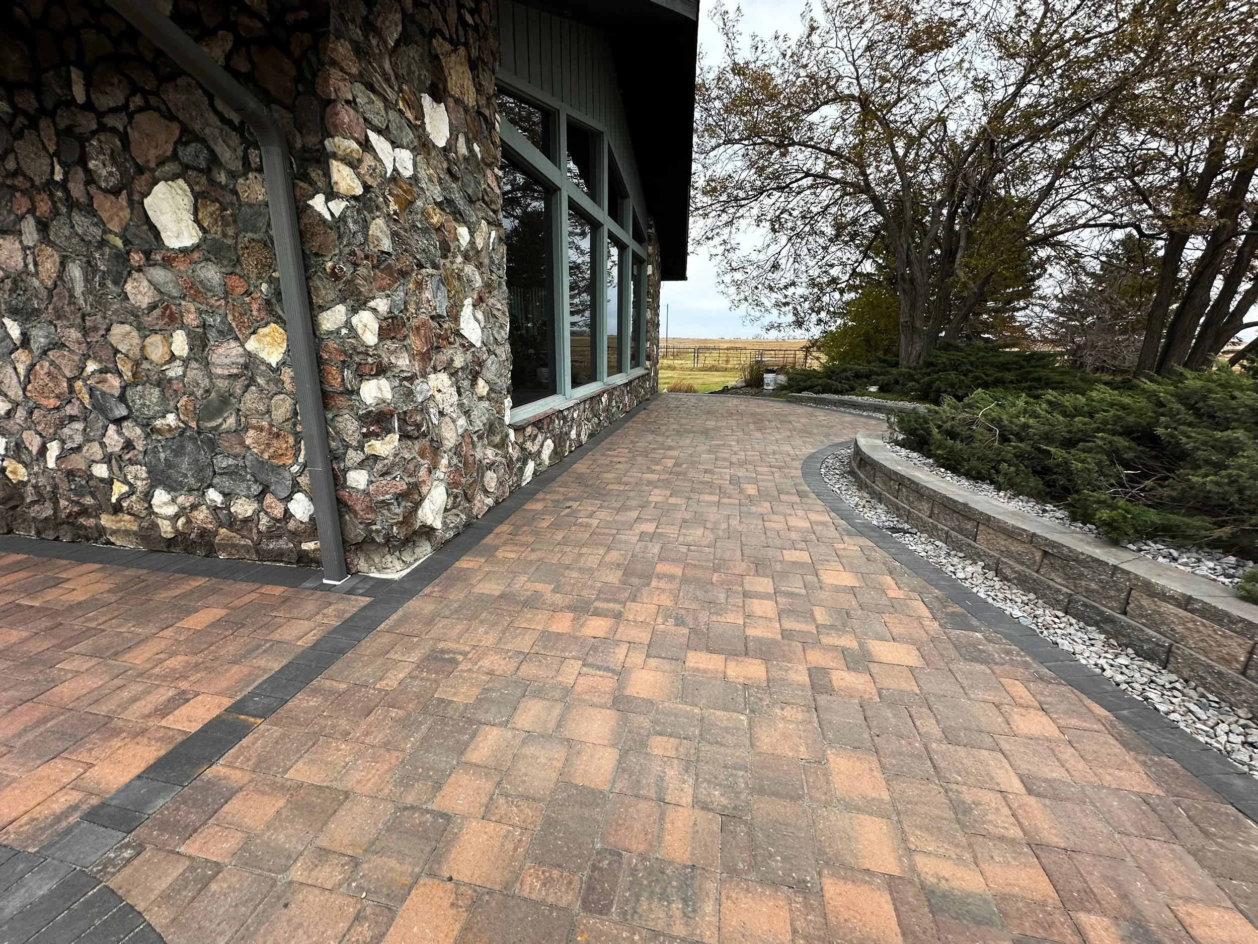 A paved brick walkway next to a stone building with large windows and a landscaped garden area with bushes and trees.