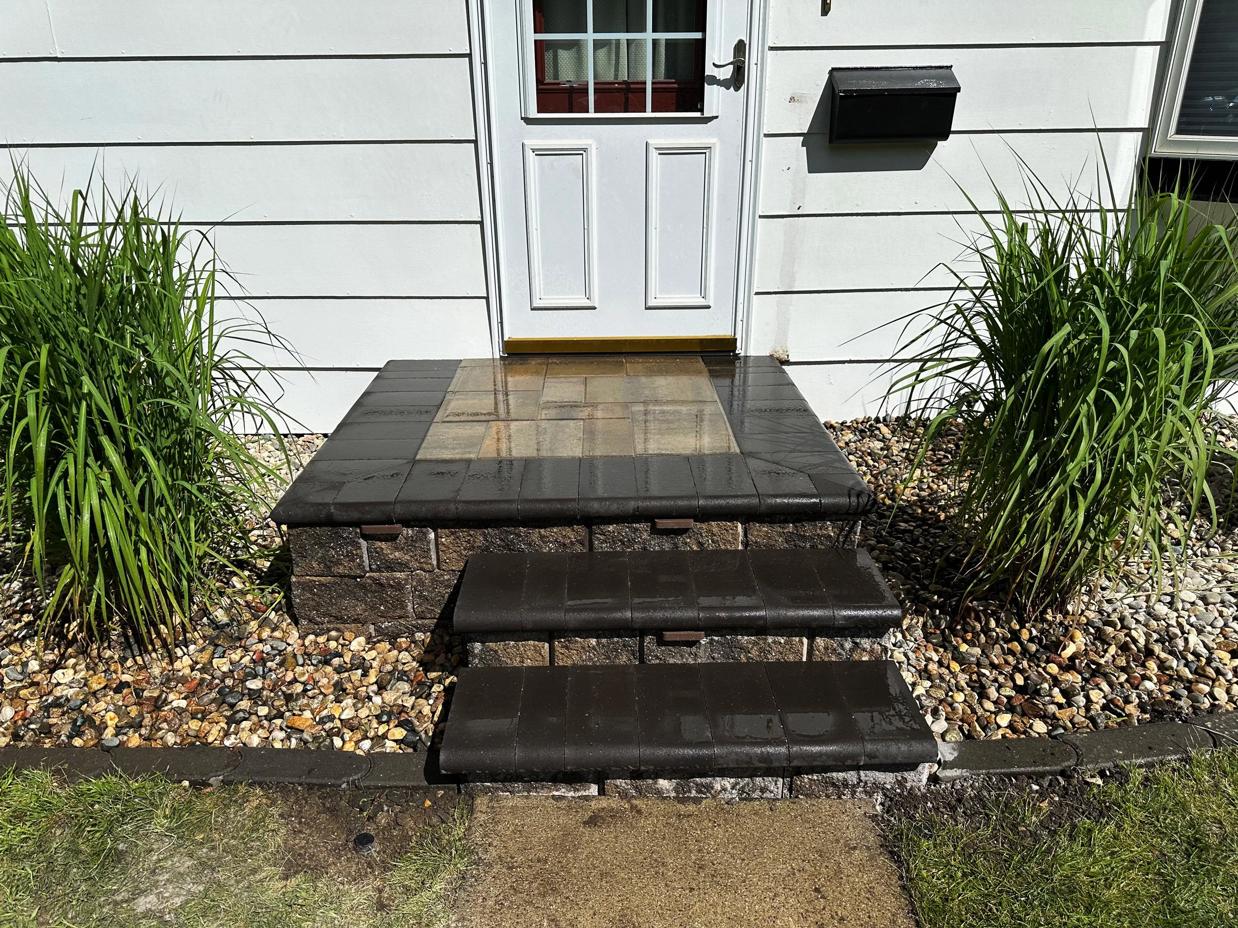 Front entrance with three dark brown steps leading up to a small concrete porch in front of a white door. The porch is bordered by plants and gravel, with a mailbox mounted on the wall to the right of the door.