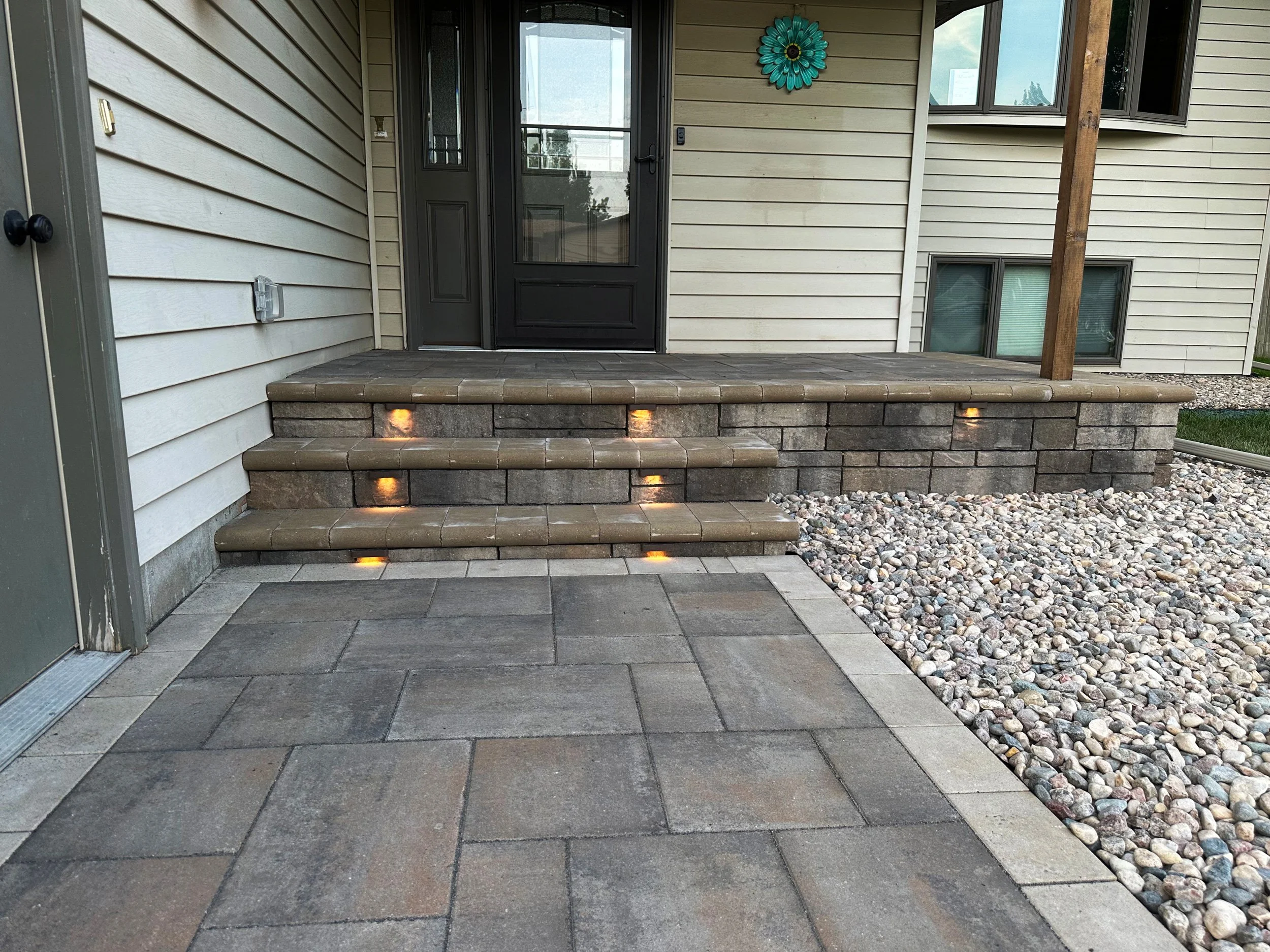 Outside of a house with a small brick porch and four stairs, with embedded lights. The porch has a black door and a window, with a decorative blue flower on the wall. The ground has stone pavers, and there are gravel and grass areas nearby.