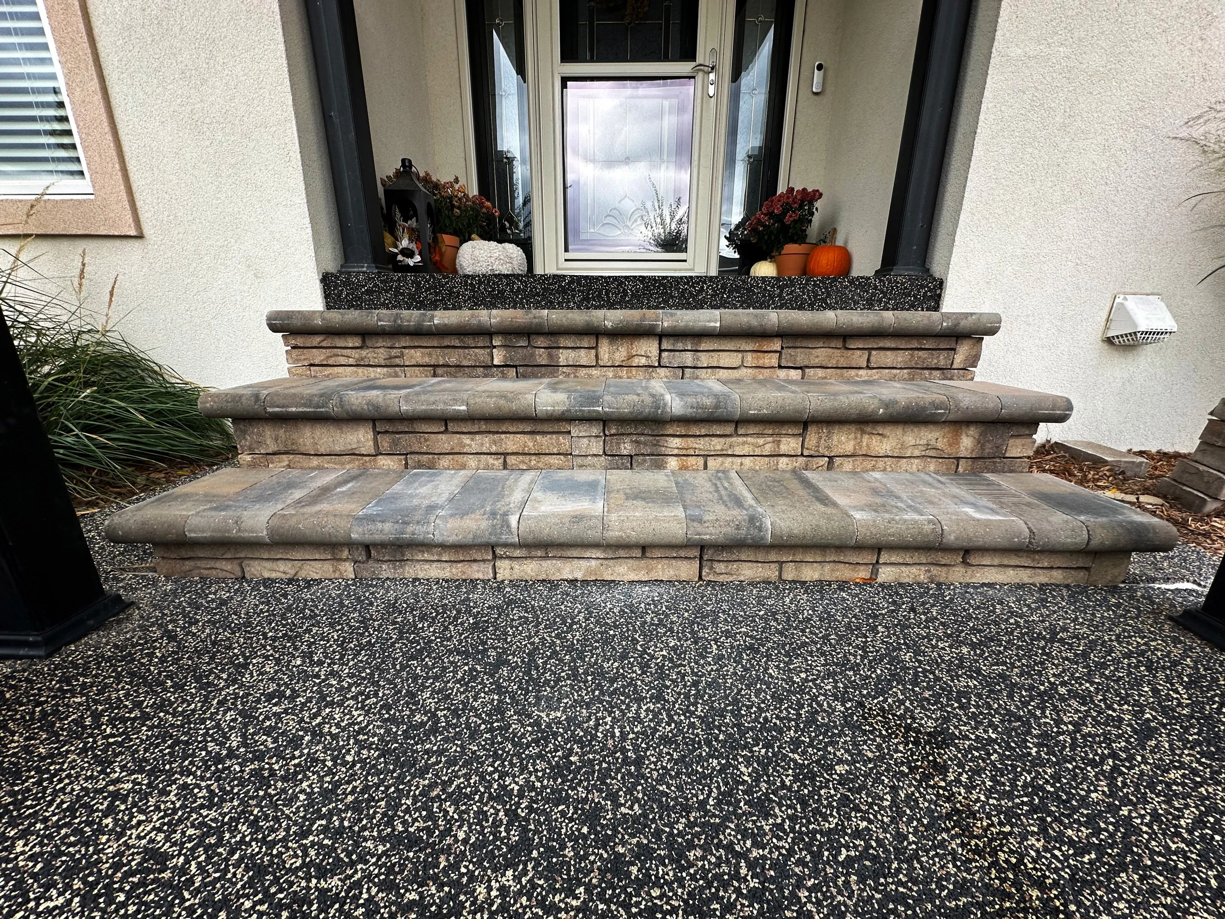 Outdoor front porch with three stone steps leading up to a front door. The steps are made of brick and stone, with black railings on each side. The porch has a textured black and white floor. Potted plants and pumpkins are placed on the porch, with a