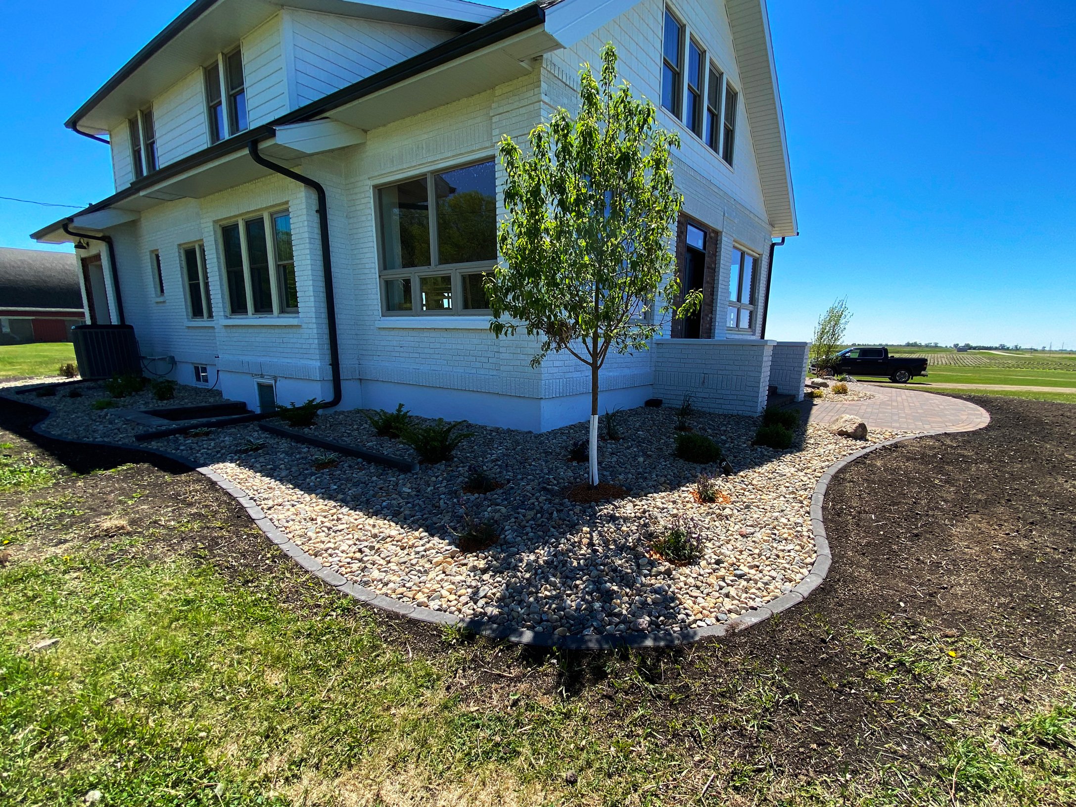 Side view of a white two-story house with a landscaped yard featuring a small tree, gravel, and a paved pathway, with a clear blue sky in the background.