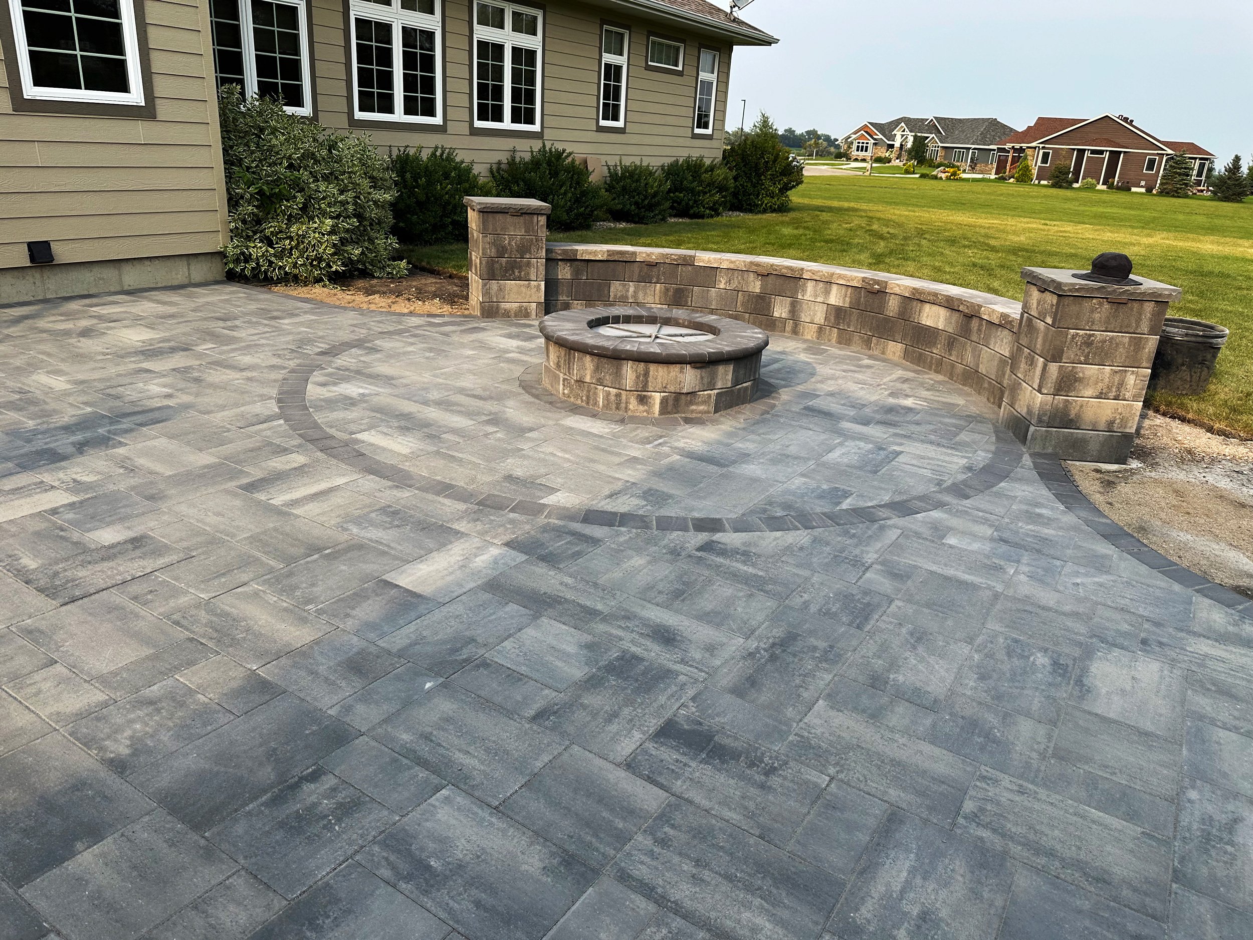 A new outdoor patio with grey paver stones, a stone fire pit, and a curved stone retaining wall, adjacent to a house with beige siding and white window frames. There is a grassy area beyond the wall and other houses in the distance.