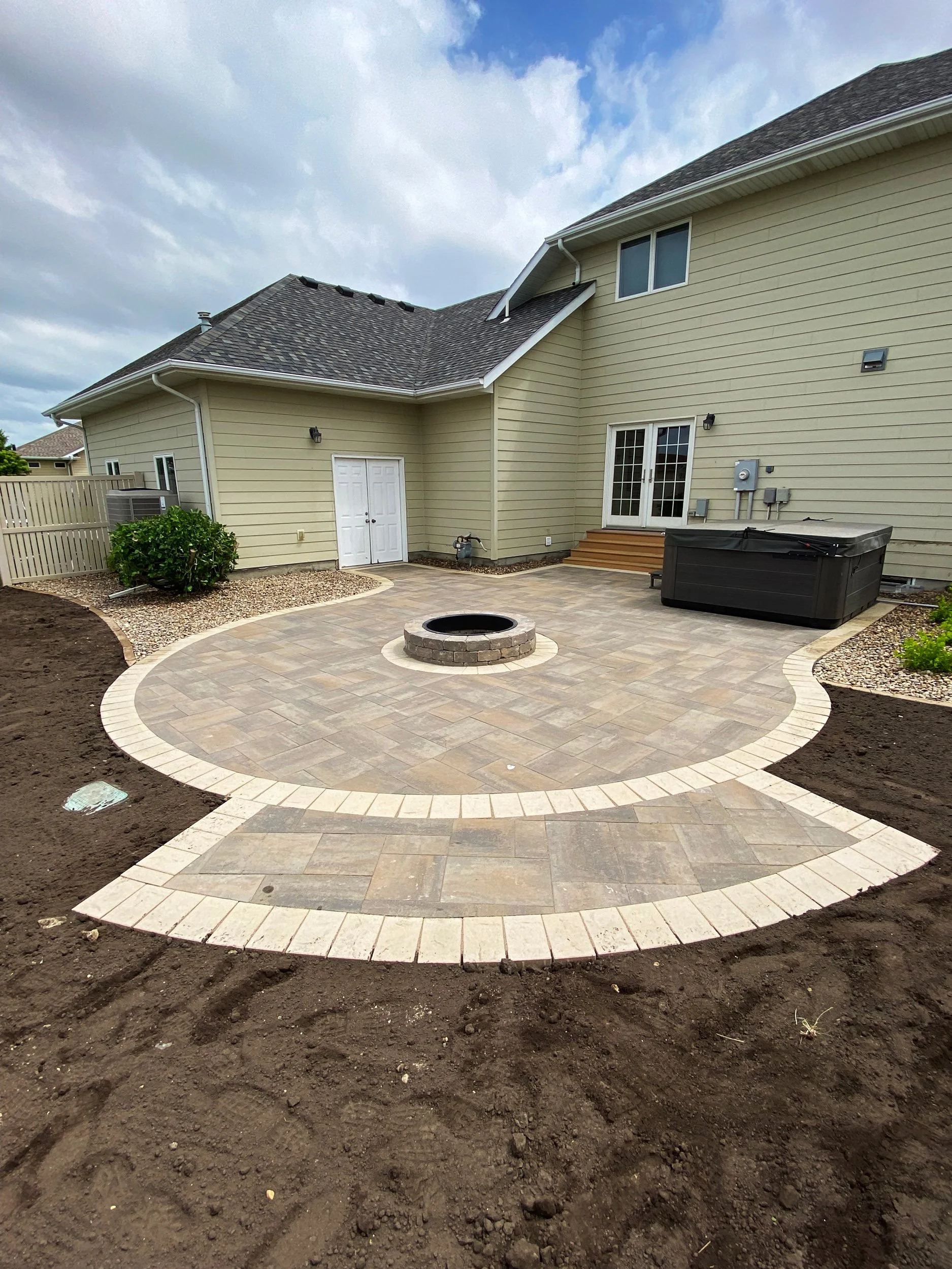 Backyard with a newly paved stone patio, a fire pit in the center, a hot tub on the right, and steps leading to a sliding door, with a beige house in the background and garden beds on the sides.
