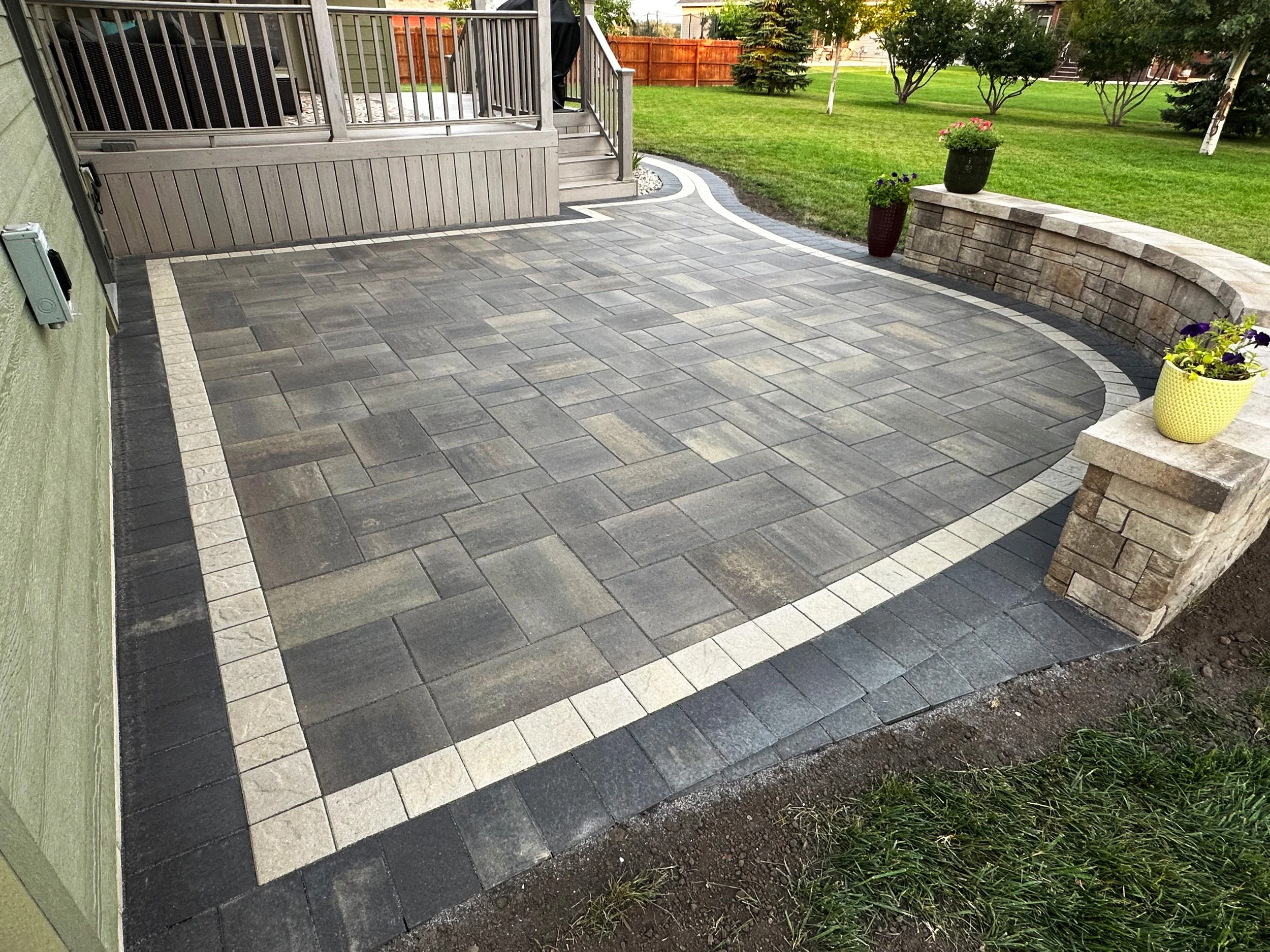 A newly paved outdoor patio area with a combination of dark gray, light gray, and black stone bricks, bordered by a white stone trim. The patio is adjacent to a green house and is decorated with potted plants on stone pillars, with a grassy lawn and 