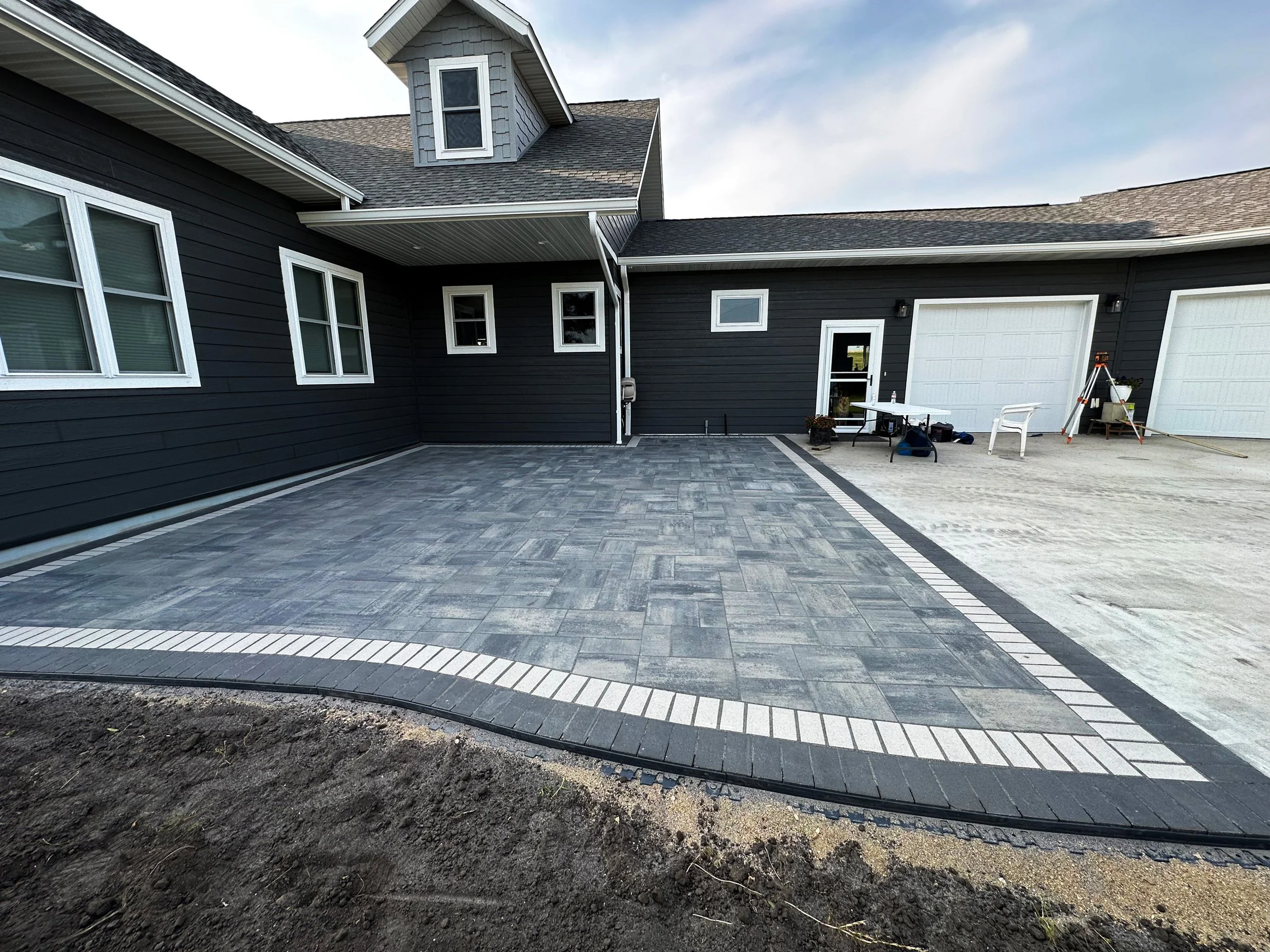 Newly paved driveway with interlocking stone pattern in front of a black house with white window frames and garage doors. Some outdoor furniture and tools are visible on the right side.