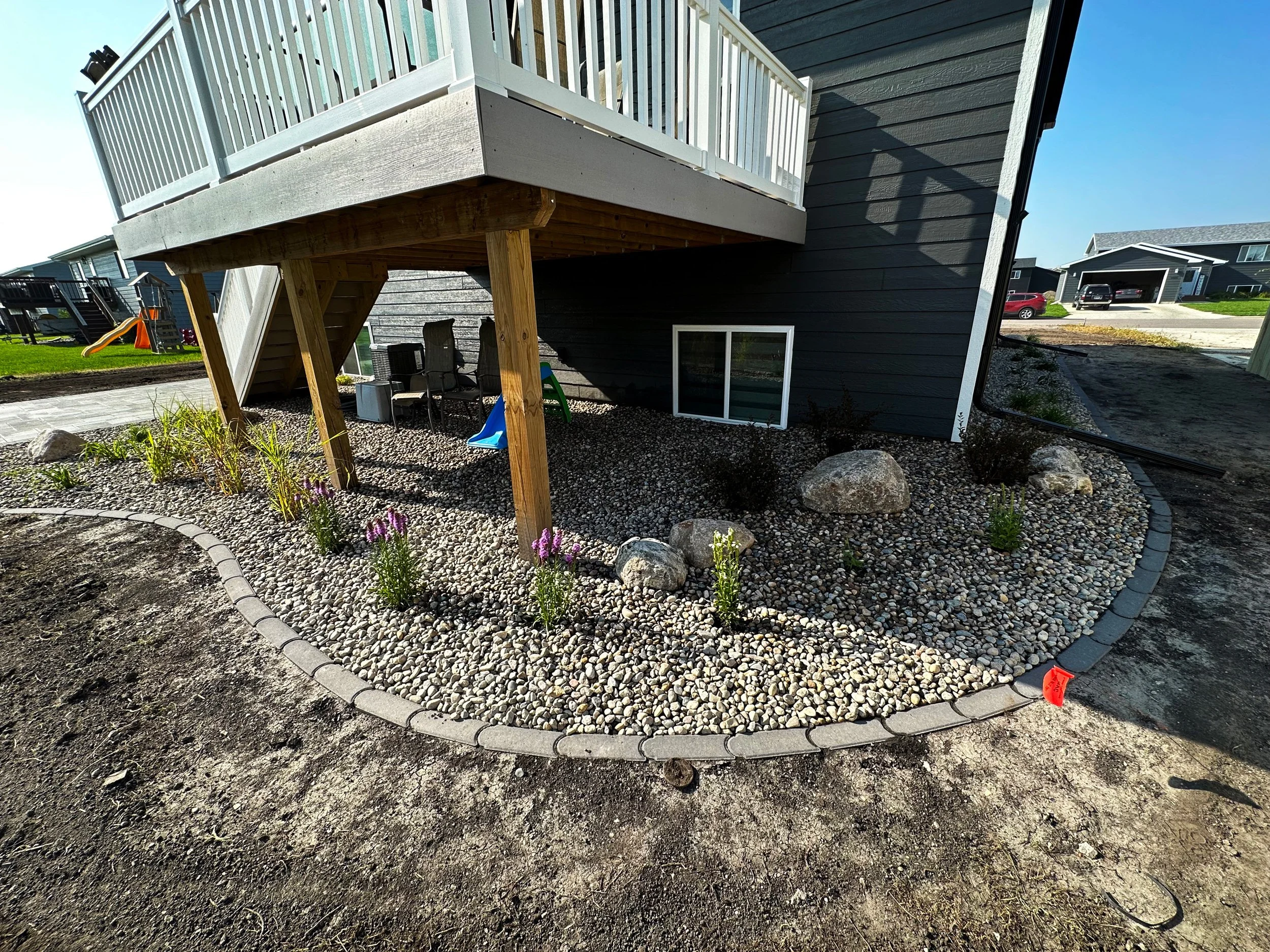 Backyard landscape with a raised deck supported by wooden posts, gravel ground cover, small purple flowers and rocks, and a surrounding black brick border.