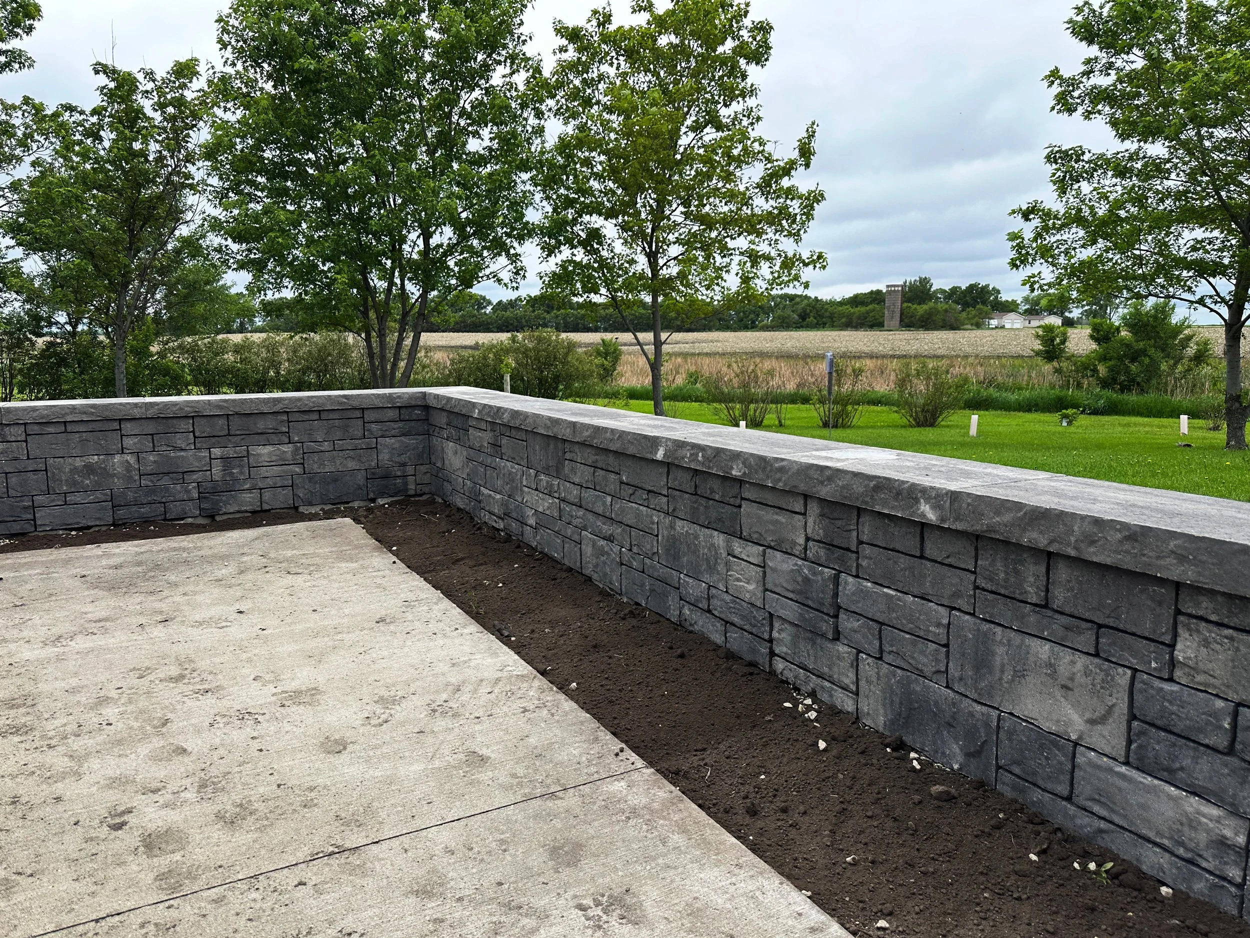 A newly constructed outdoor patio with a gray stone wall and concrete flooring, overlooking a grassy yard with trees and open fields in the distance.