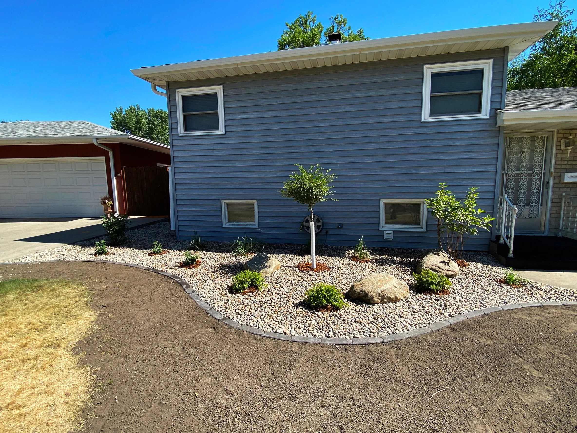 Front yard of a house with a landscaped garden bed filled with small rocks, bushes, plants, and large rocks, with a small tree in the center, and a blue house with white trim and a white staircase on the right.