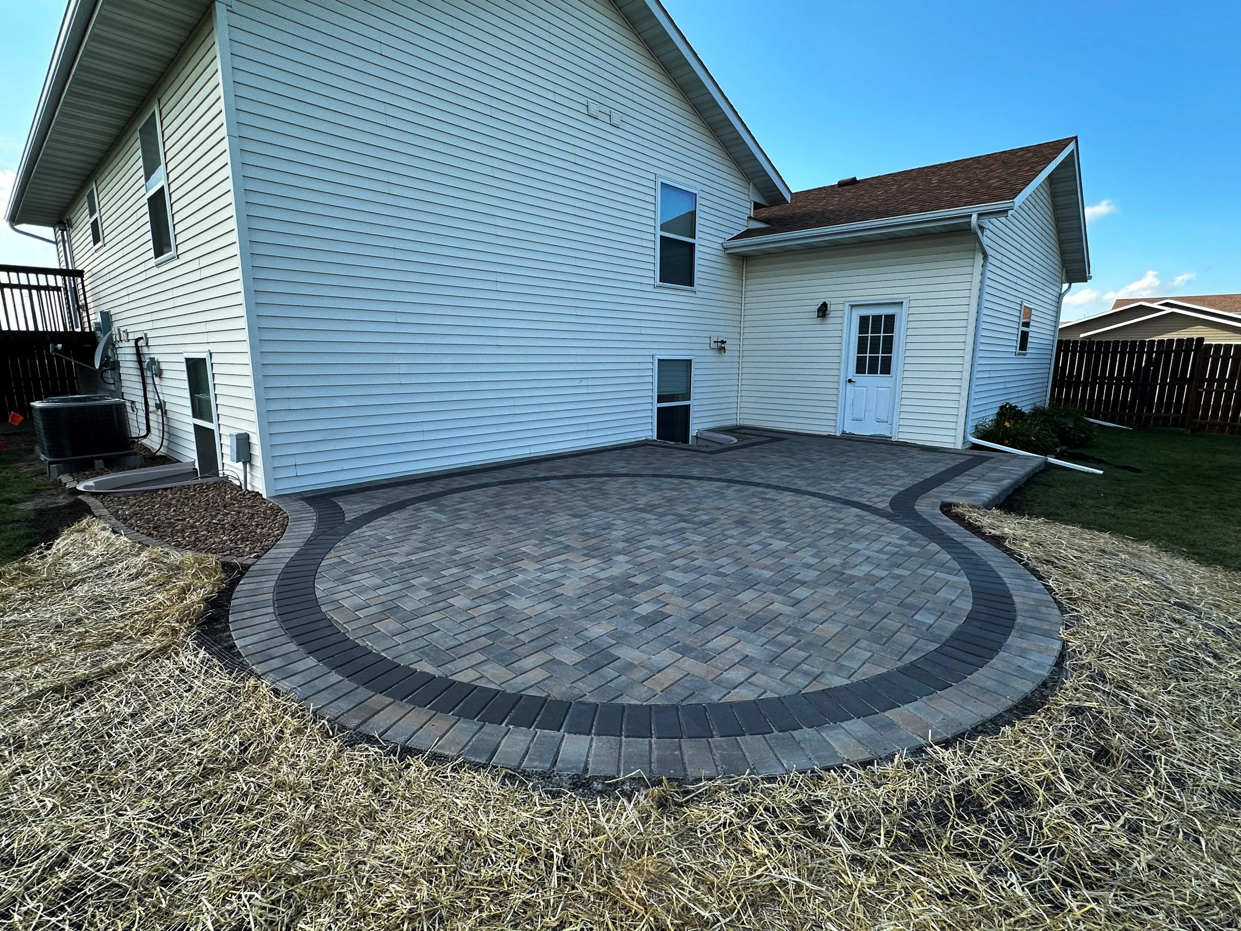 Backyard patio with interlocking pavers in front of a house with white siding, a door, and windows, bordered by straw and mulch.