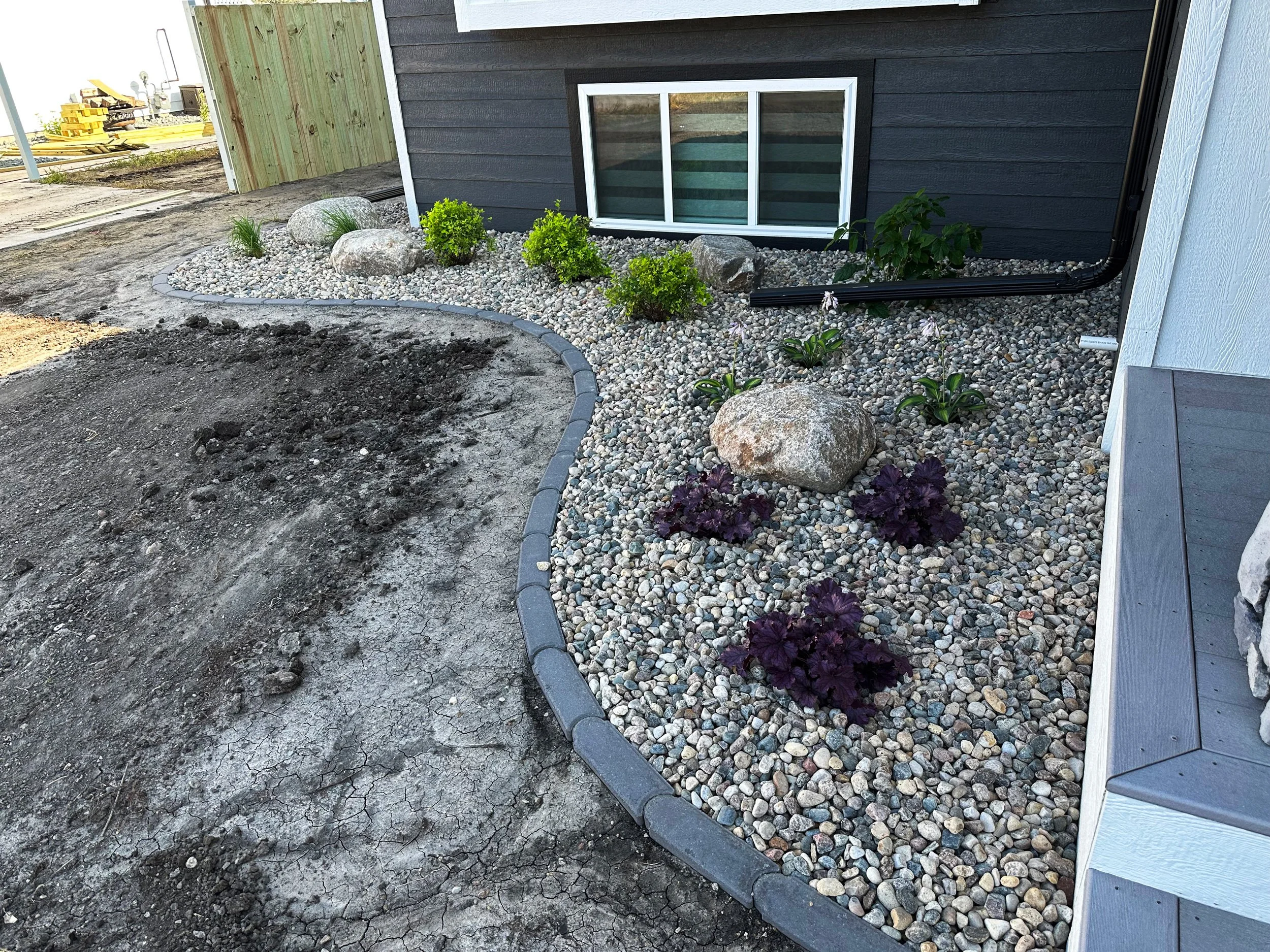 Newly landscaped backyard with a curved border of edging stones, filled with multicolored small rocks and garden plants, alongside a white and black house wall and window. There is a section of disturbed earth and dirt in the foreground, with constru