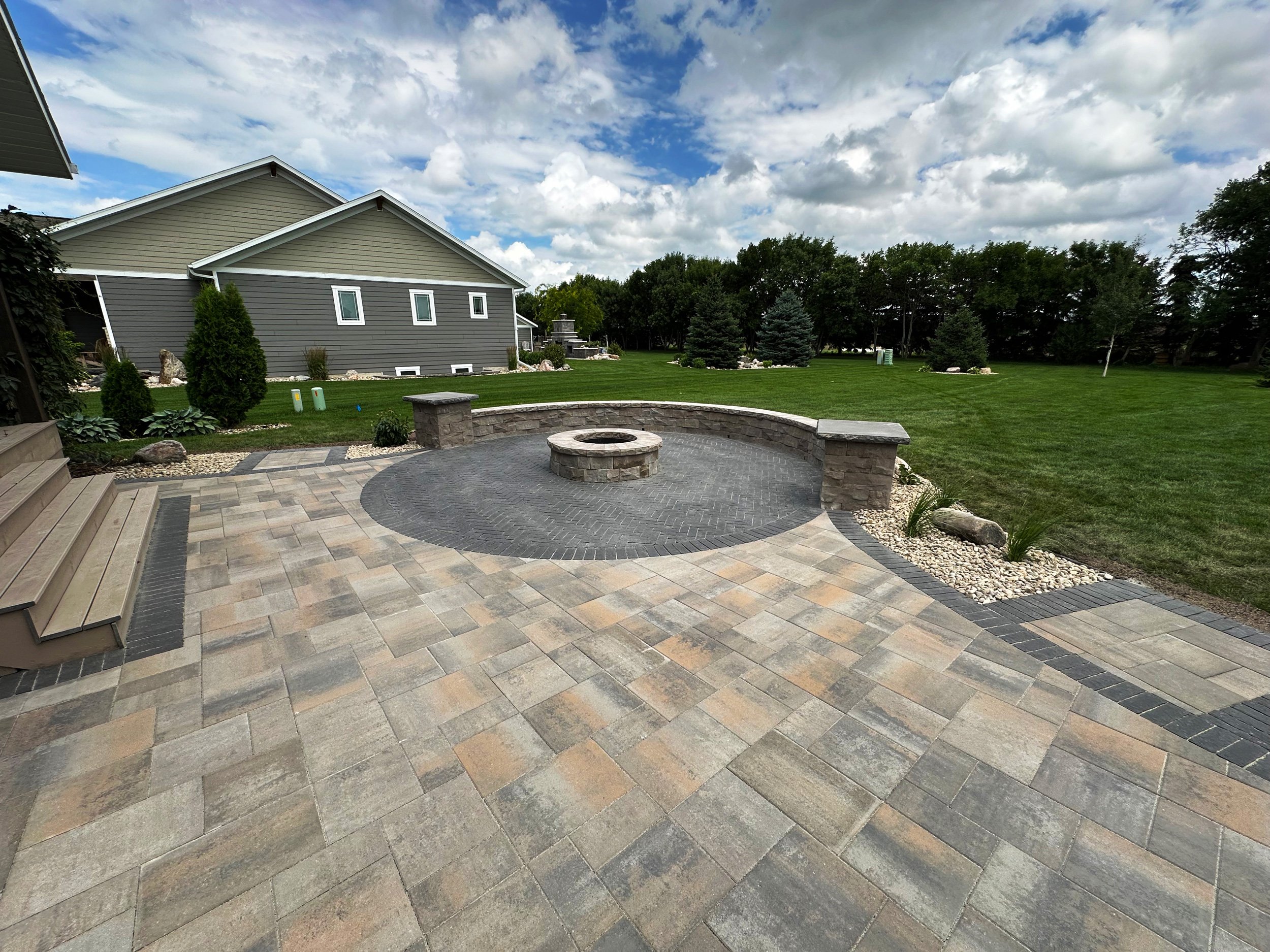 Backyard patio with stone pavers, seating area with stone pillars, and a fire pit, surrounded by green lawn and trees under a partly cloudy sky.
