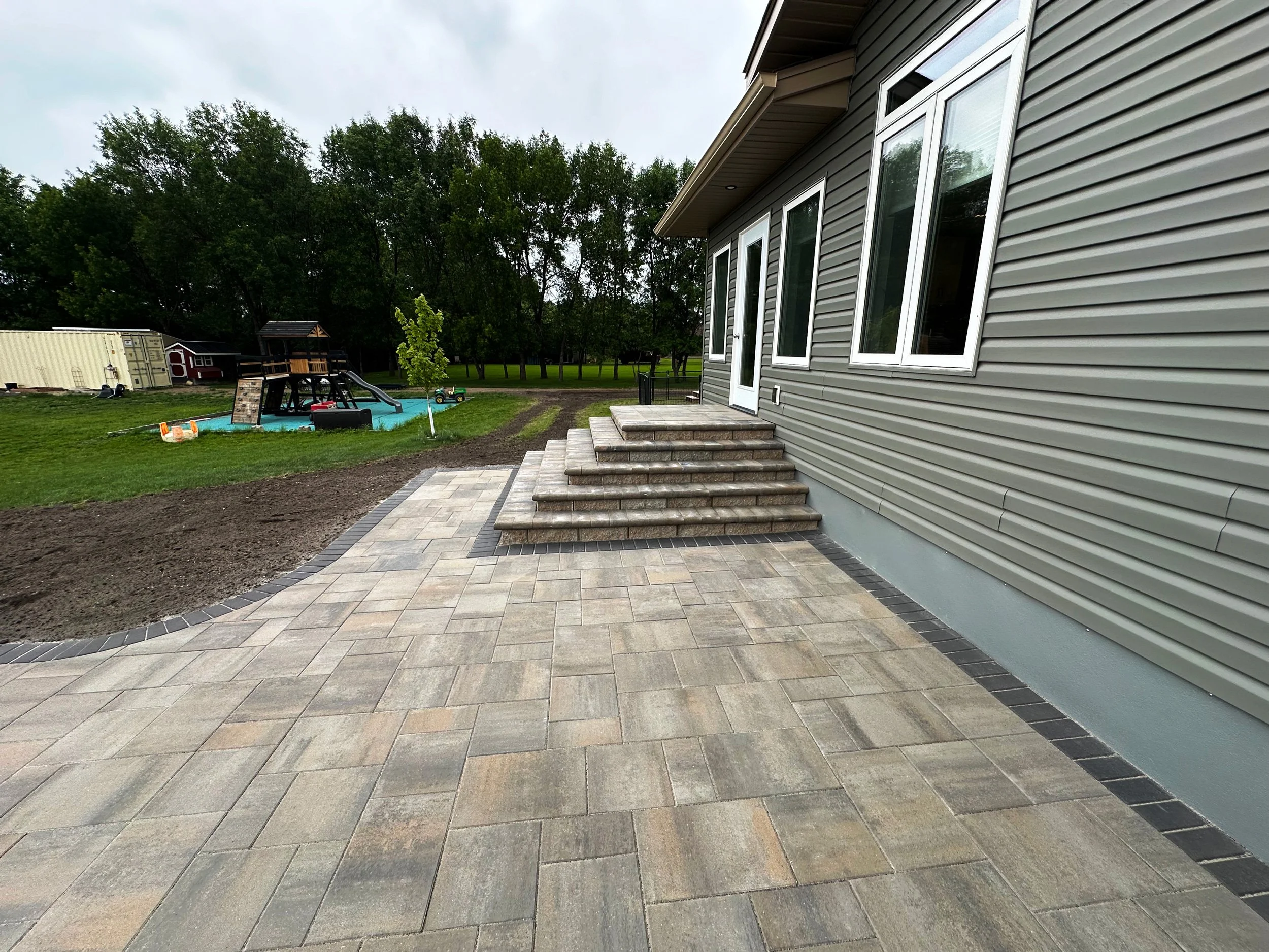 Backyard patio with gray stone stairs and new paver walkway adjacent to gray siding house, overlooking a grassy yard with a children's play area featuring a wooden playset and slide, and a forested area in the background.