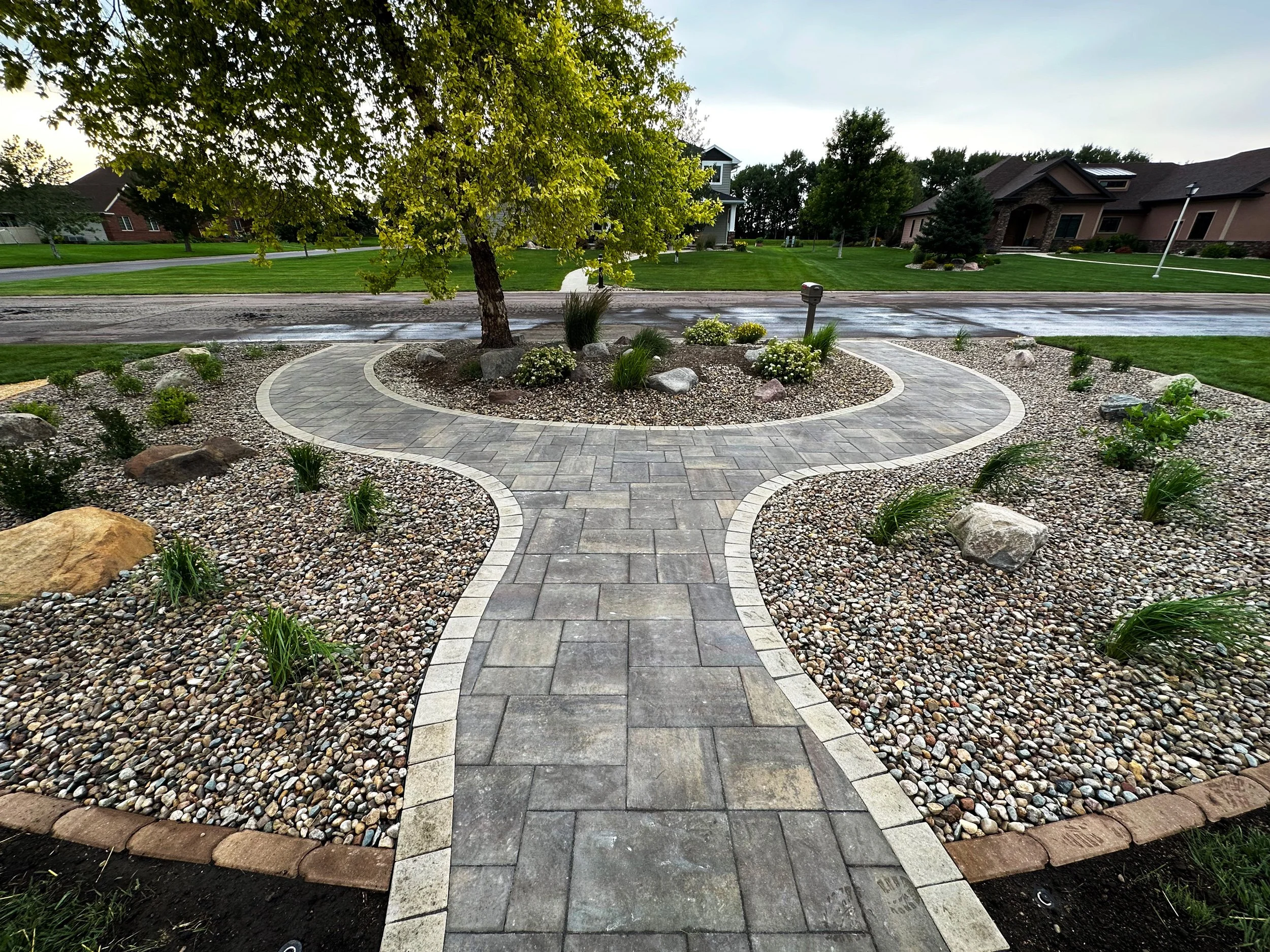A backyard garden with a winding brick pathway, a small tree, and landscaped rocks and plants, with houses and a street in the background.